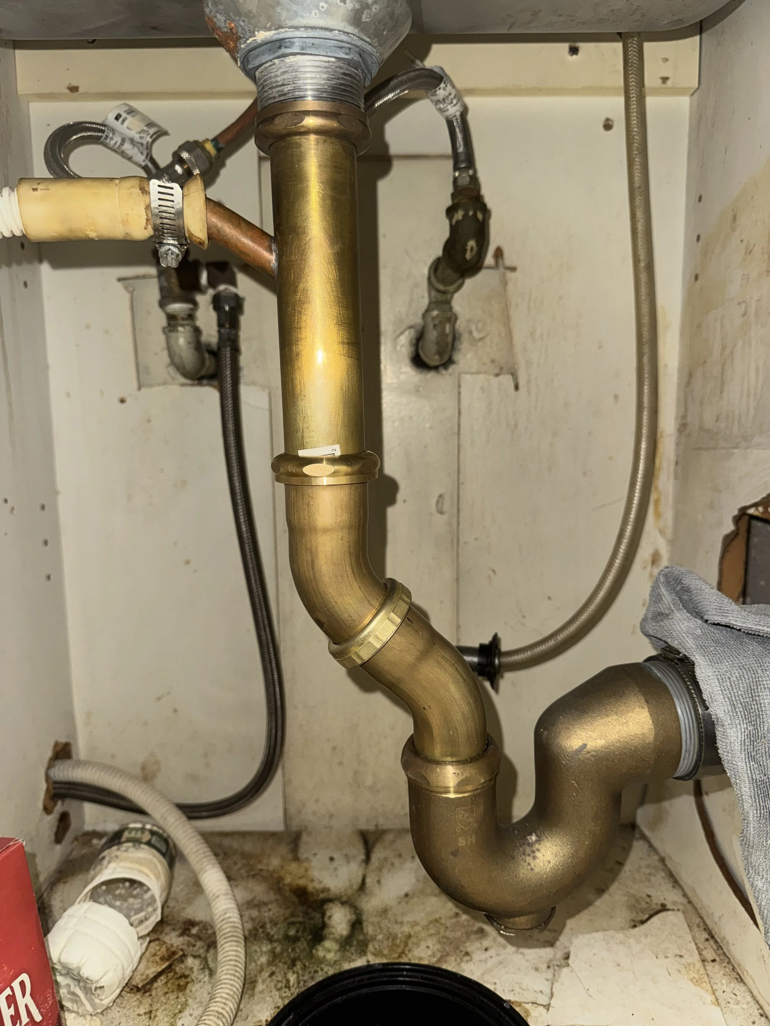 Underground plumbing pipes, including a large brass pipe with a bend, connected to smaller pipes and hoses under a kitchen or bathroom sink.