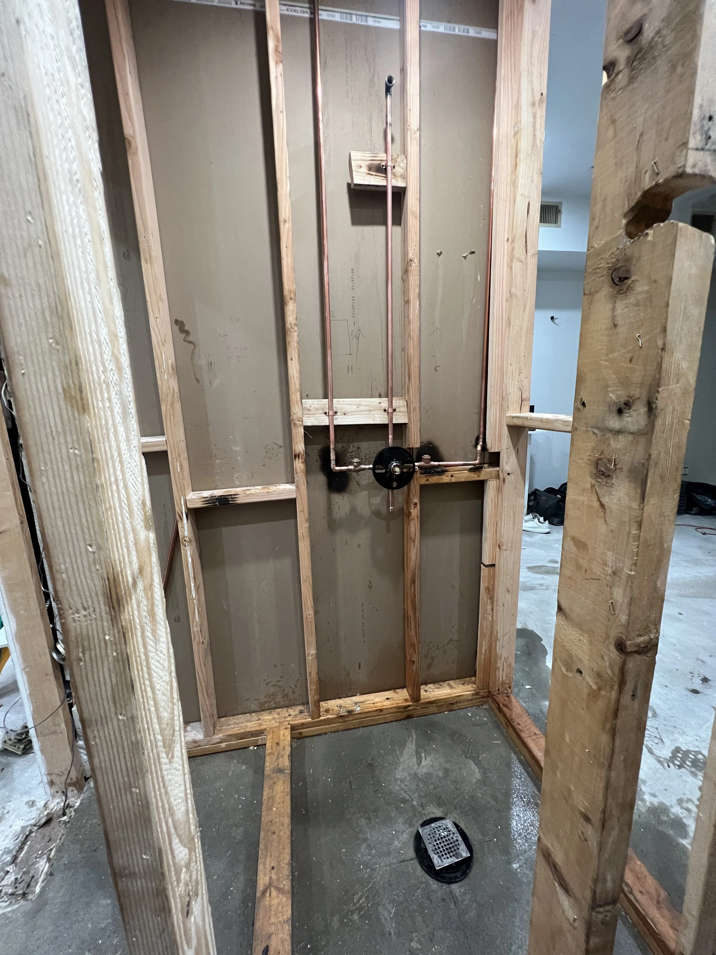 A bathroom in the process of being renovated with exposed wooden framing and plumbing for a toilet. The floor is concrete with a drain cover visible.