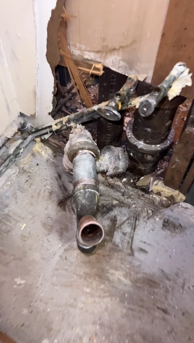 A close-up of a pipe under repair in a construction or plumbing setting, with pipes, fittings, and a hole in the wall surrounded by debris and exposed wood.