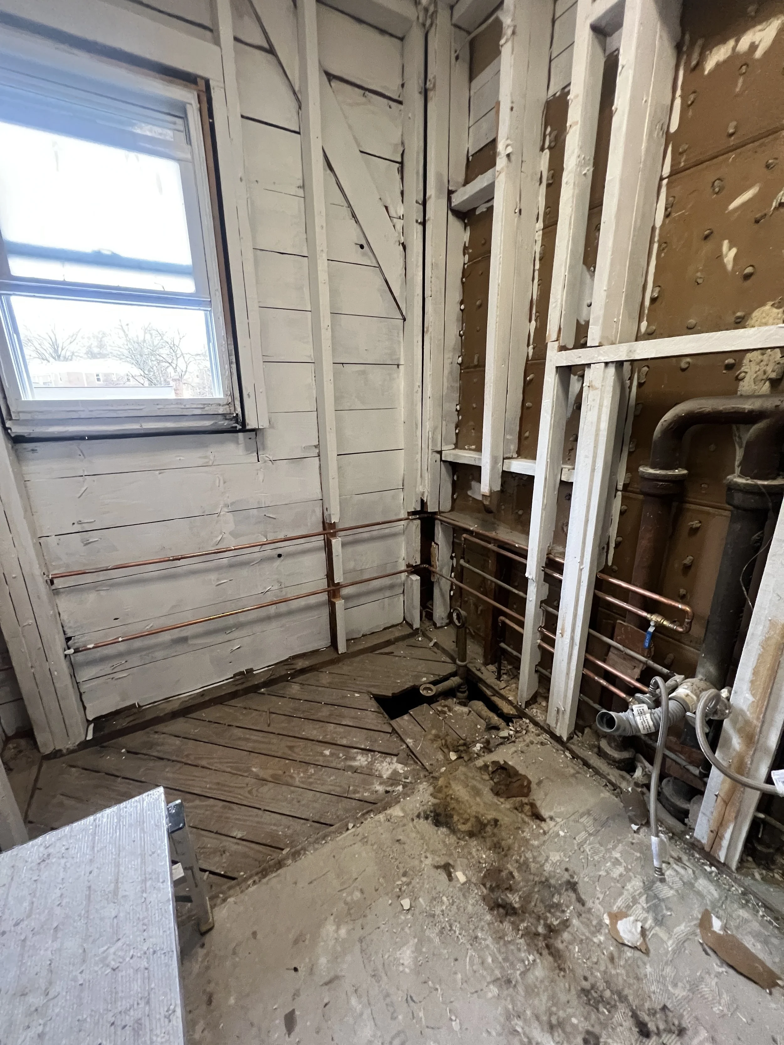 Room under renovation with exposed brick wall, copper plumbing pipes, and stripped wooden floor.