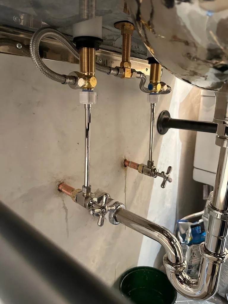 Underneath a sink showing plumbing fixtures including copper pipes, brass fittings, and flexible hoses connected to the sink drain and water supply lines.