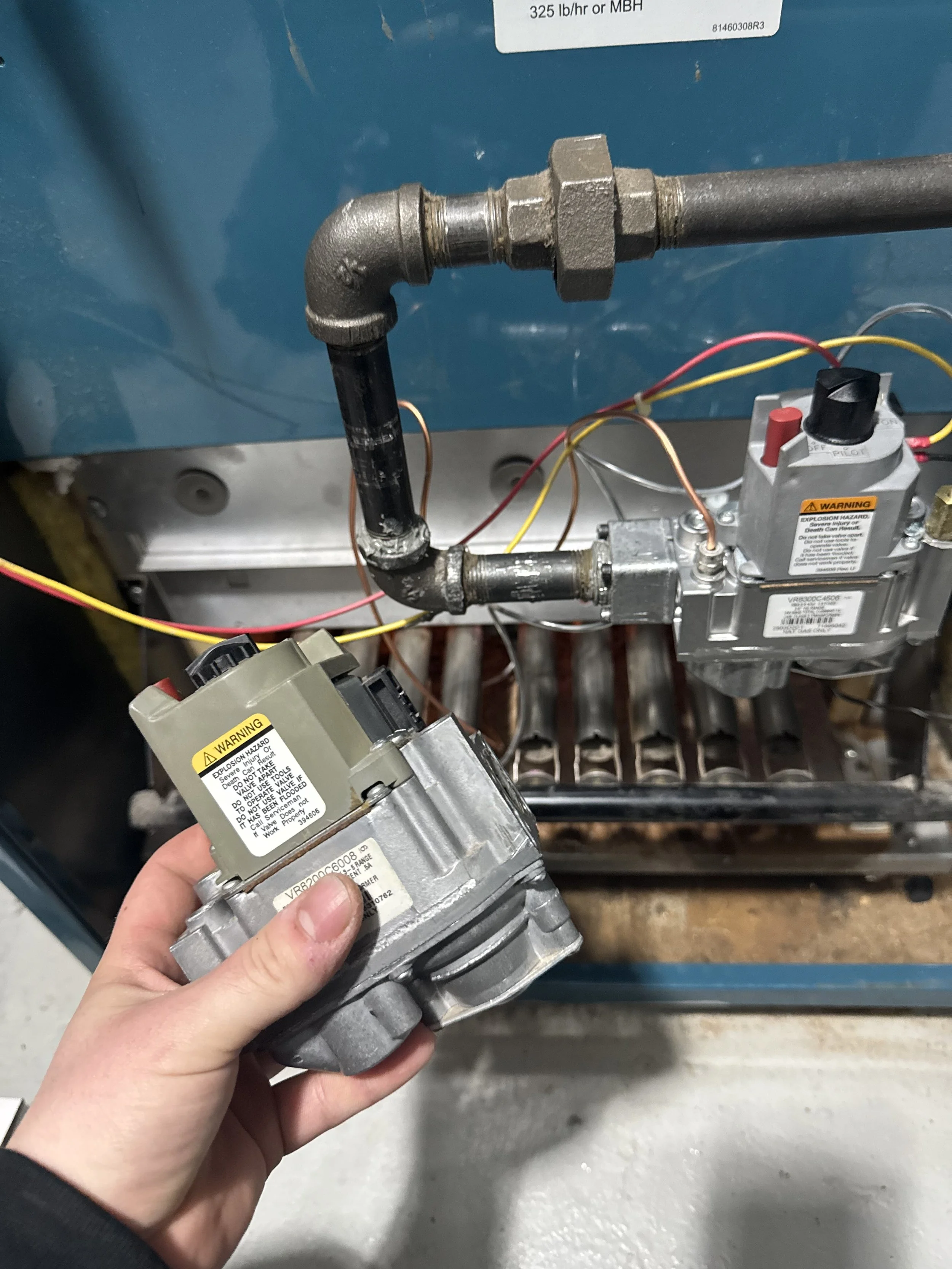 A person holding an HVAC component near the inside of an air conditioning unit, with pipes, wires, and electrical components visible.