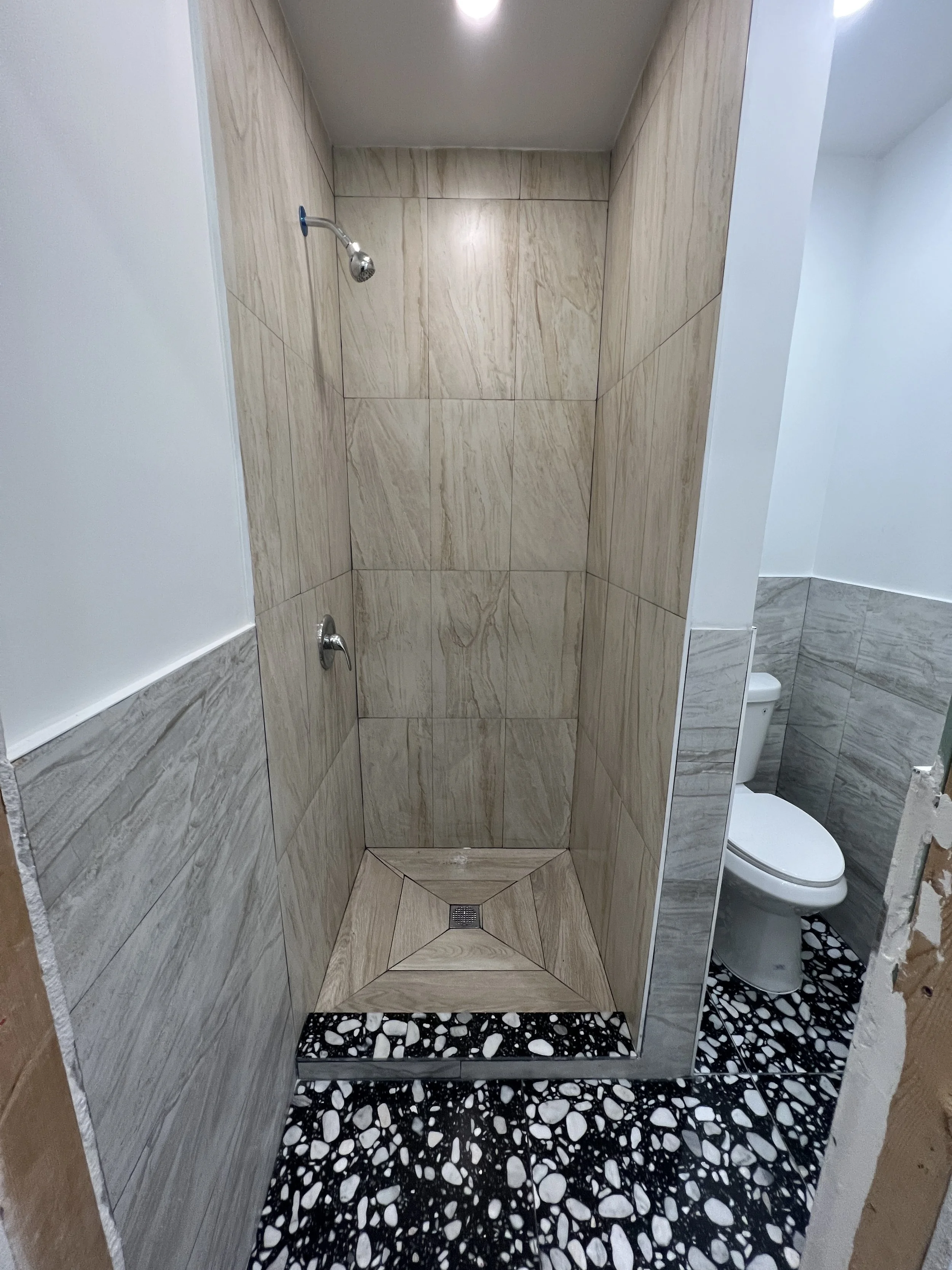 A walk-in shower with beige marble tiles and a square drain, adjacent to a toilet in a small bathroom with patterned black and white floor tiles.