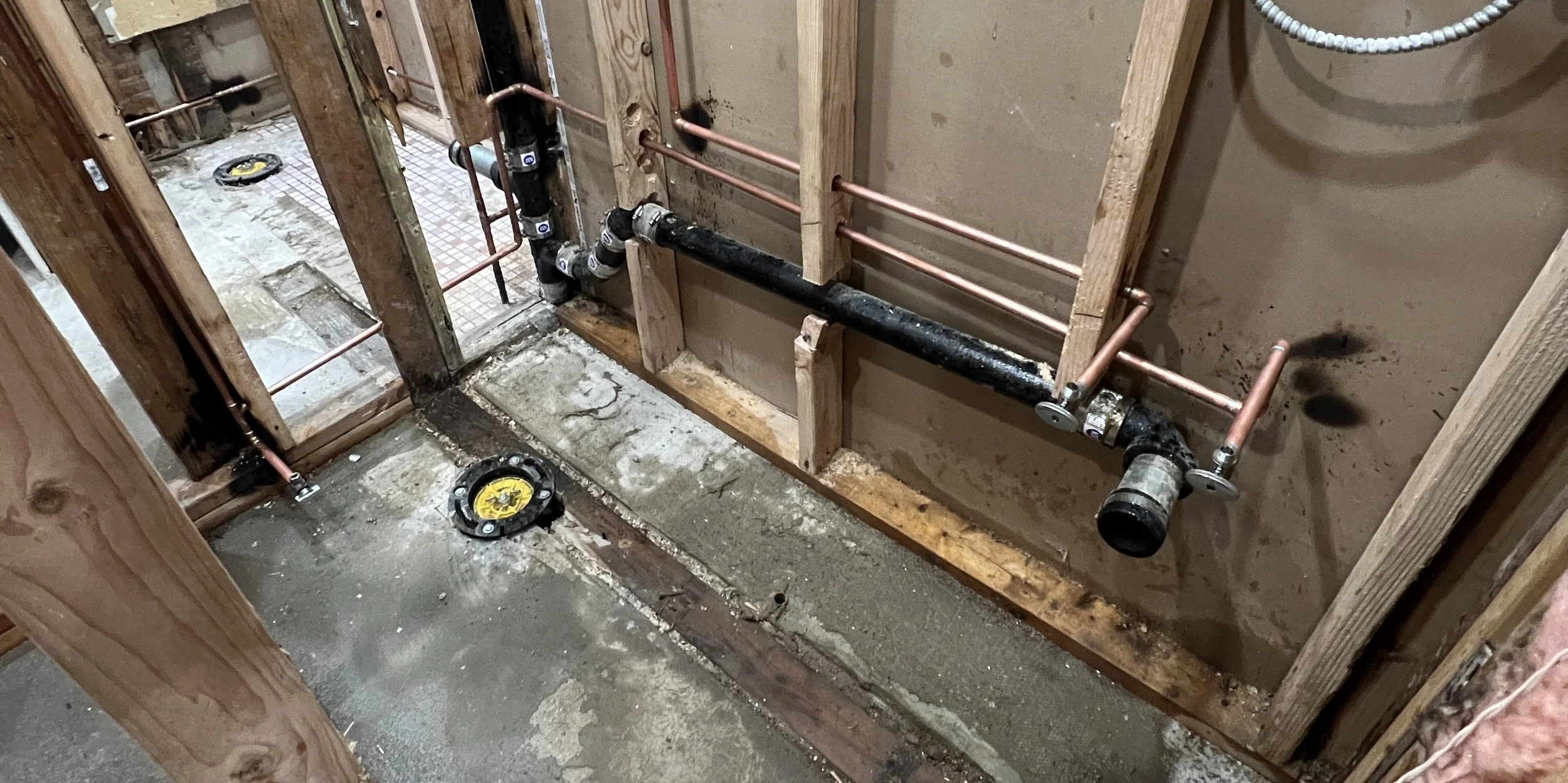Plumbing installation in a building under construction, showing black and copper pipes, and a wall with exposed wooden framing.