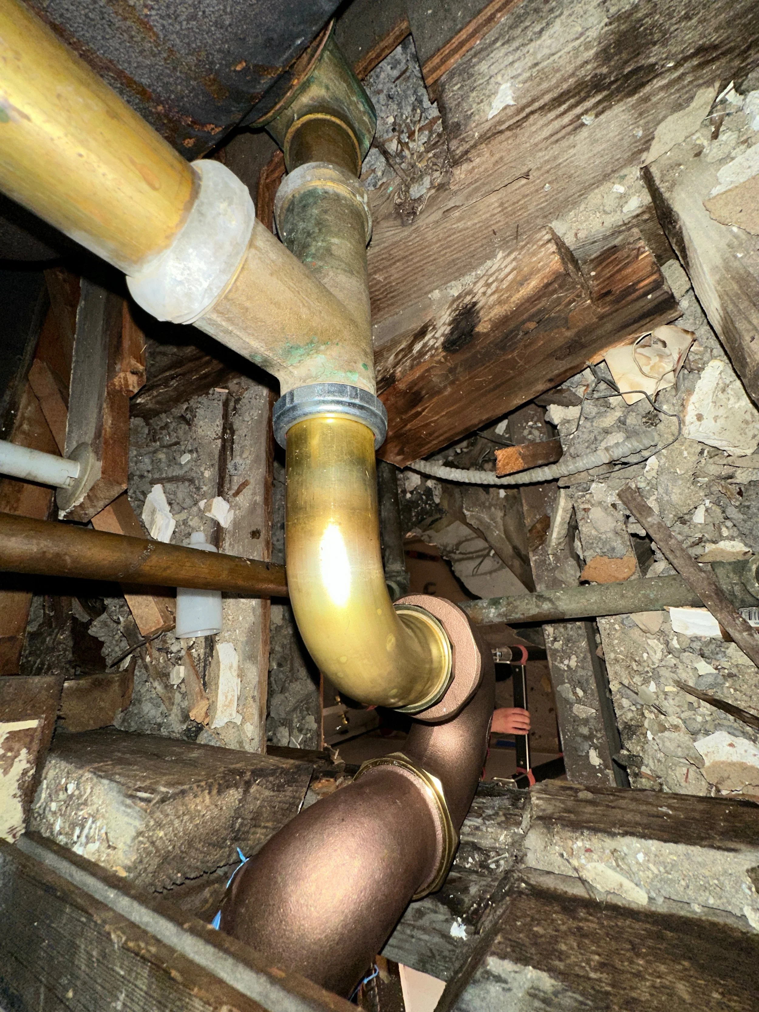 Close-up of plumbing pipes, including a yellow pipe and a bronze pipe, surrounded by construction debris and wooden framing in an unfinished space.