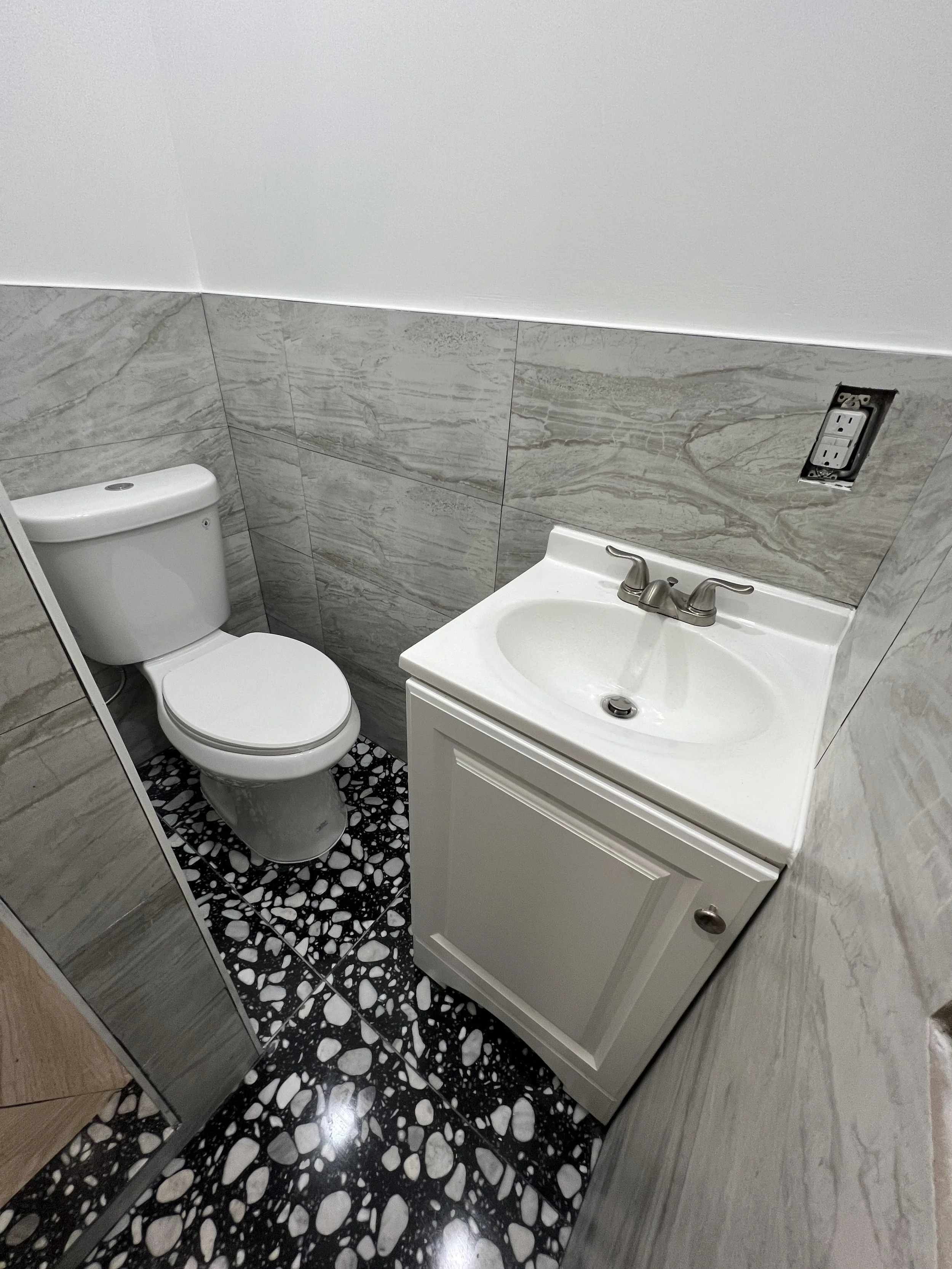 Small bathroom with a white toilet, a white sink with a gray faucet, a gray marble tiled wall, and black and white pebble patterned flooring.