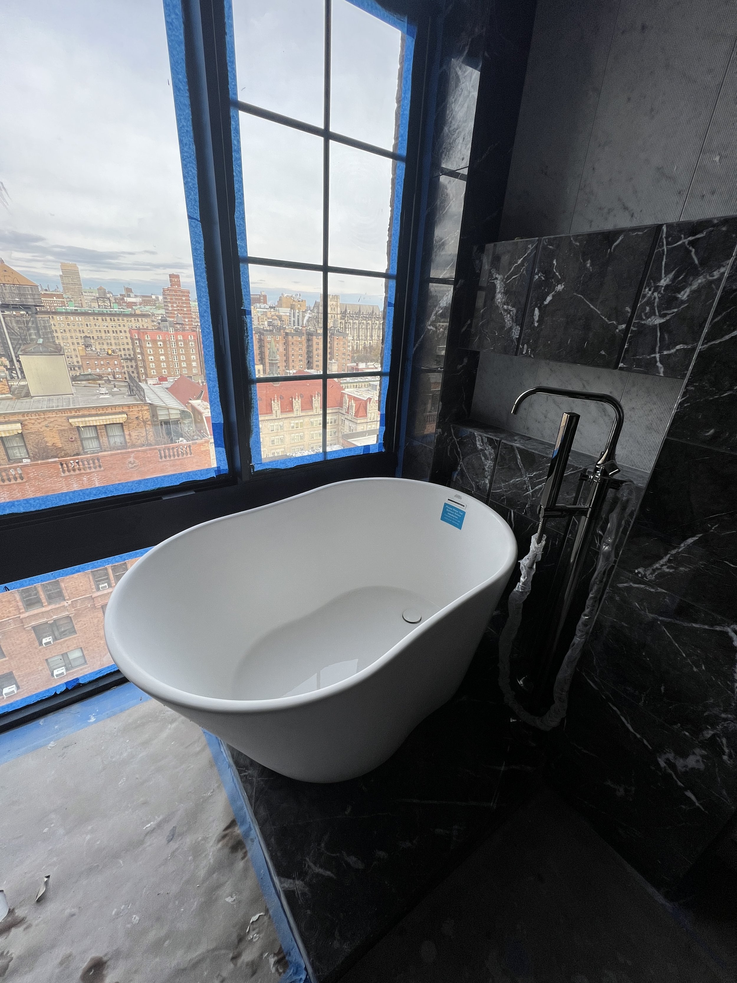 Modern bathroom with a freestanding white bathtub near large window showing city skyline, black marble walls, and construction materials on the floor.