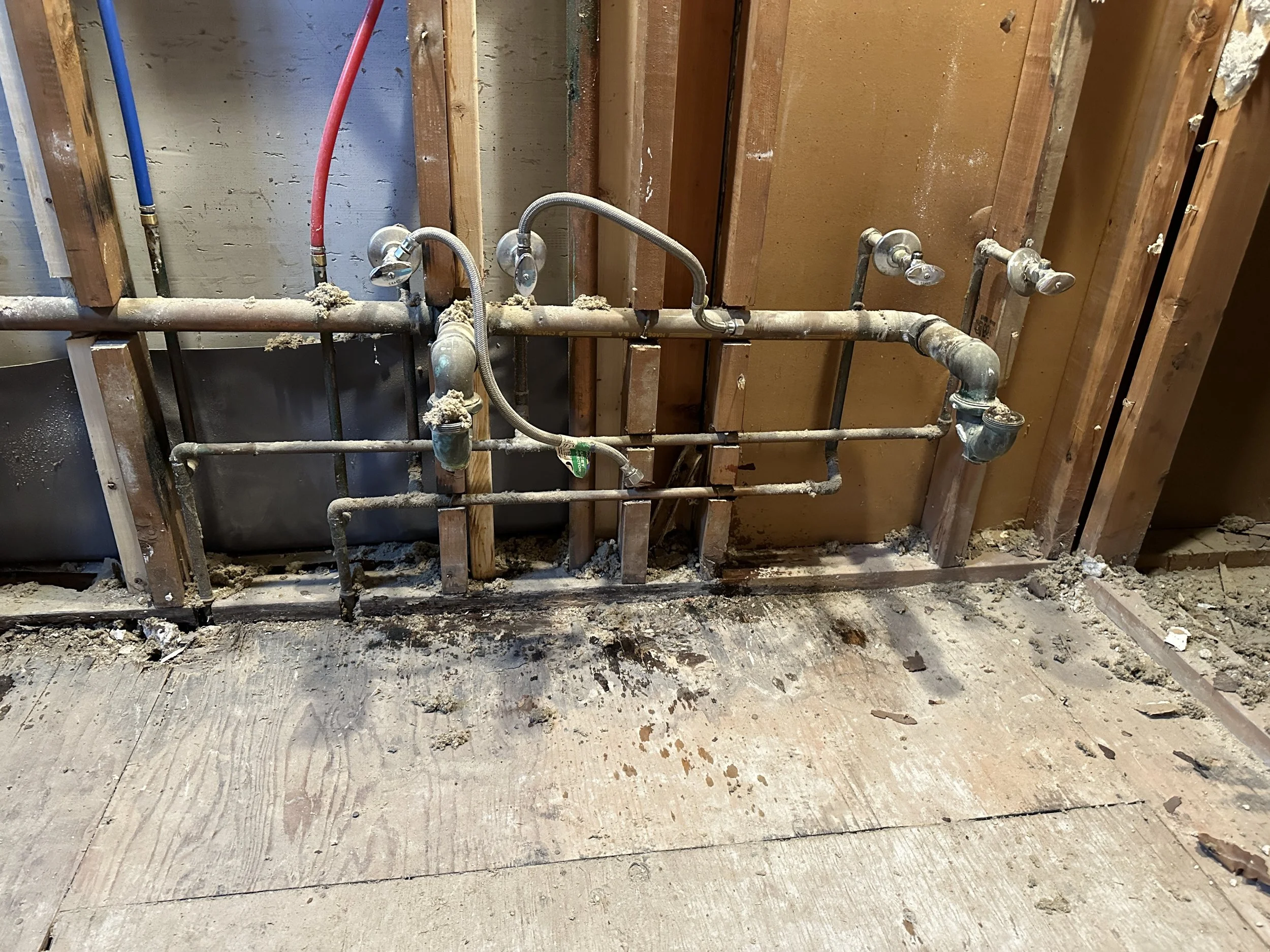 Exposed plumbing pipes and fittings in a partially demolished wall, with visible wooden studs and debris on the floor.
