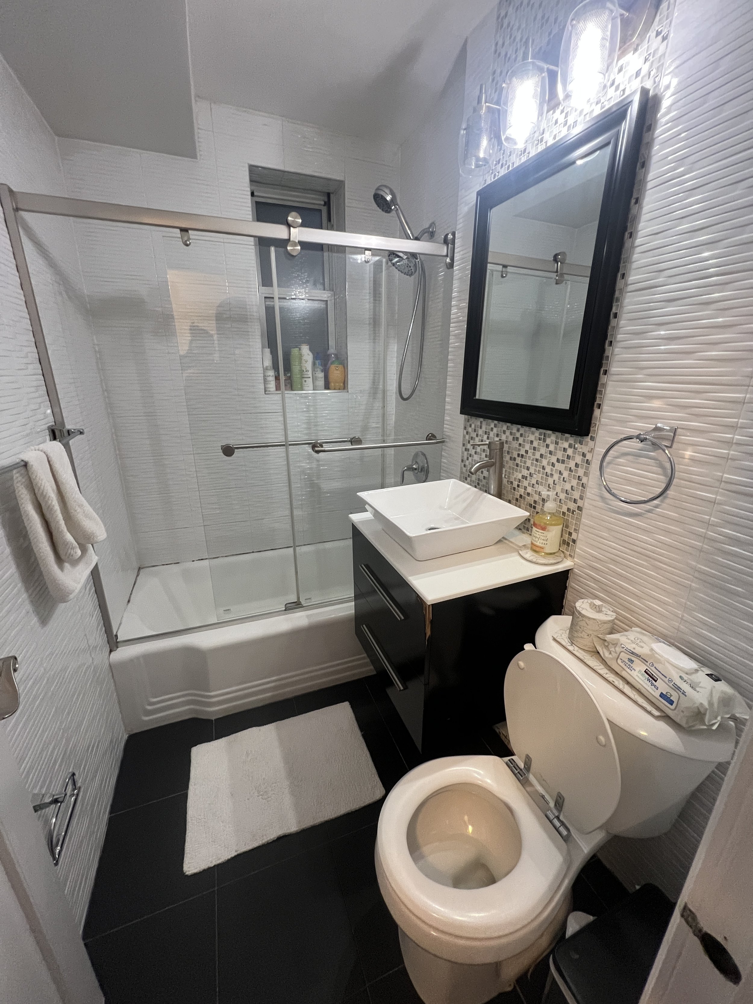 A small modern bathroom with a glass shower stall, a white sink on a black vanity, a large black-framed mirror, a toilet with a package of wipes on top, and white textured wall tiles.