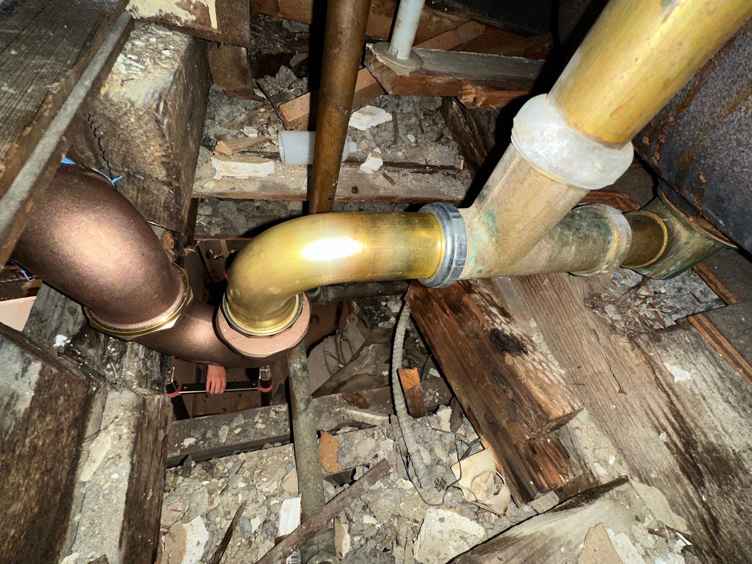 Close-up of plumbing pipes, including a copper pipe and a brass pipe, in an attic with wooden beams, debris, and insulation material.