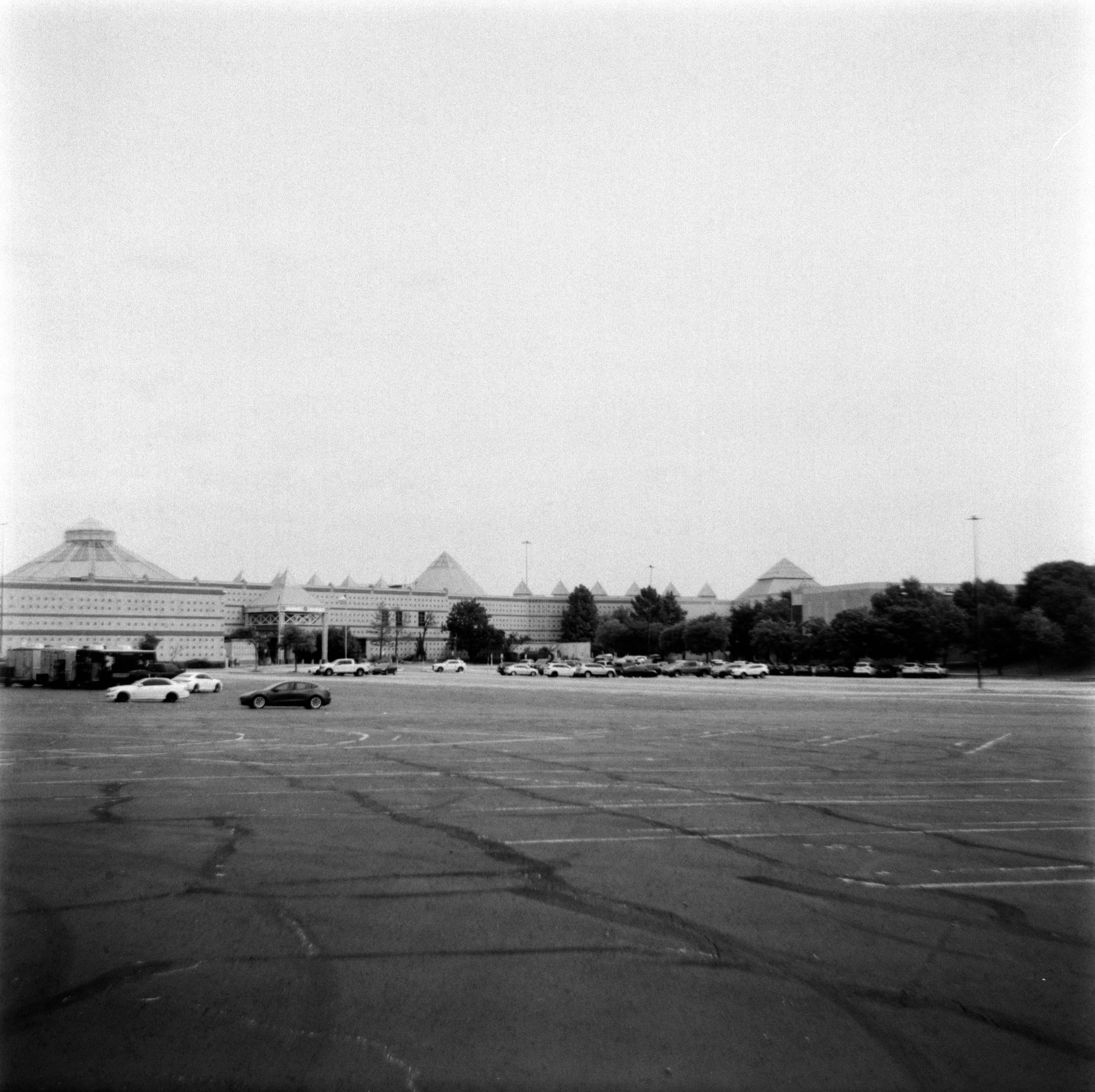 An expansive parking lot with scattered cars, with a large building featuring pyramid-shaped roofs and modern architecture in the background, under a clear sky.