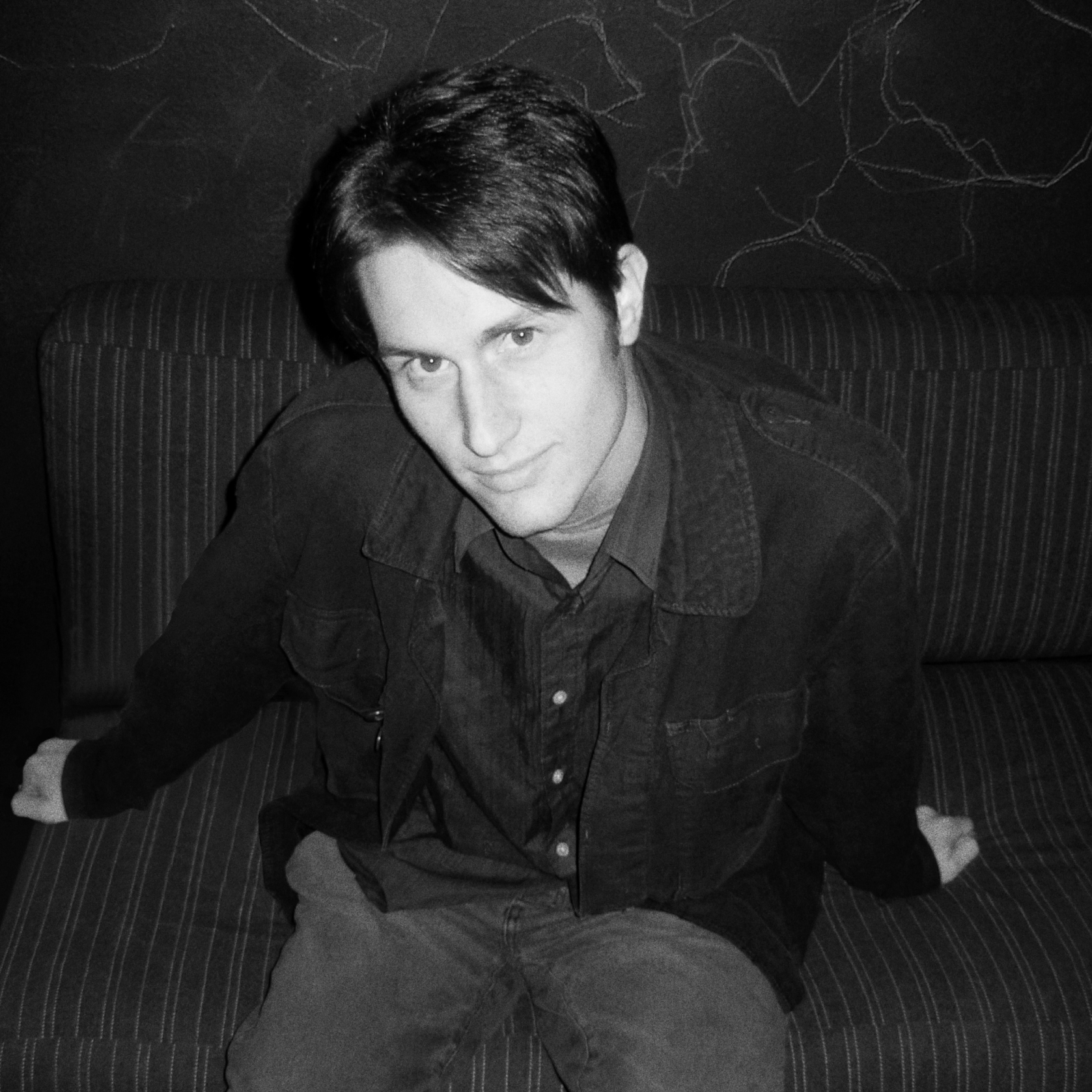 A young man sitting on a striped couch in black and white, looking at the camera with a slight smile, wearing a dark denim jacket over a shirt.