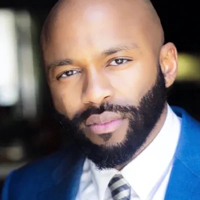 a professional close-up headshot of black man with a shaved head and a neatly groomed full beared. he is wearing a blue suit jacket, a white dress shirt, and a silver-and-black striped tie, looking directly at the camera with a confident expression.