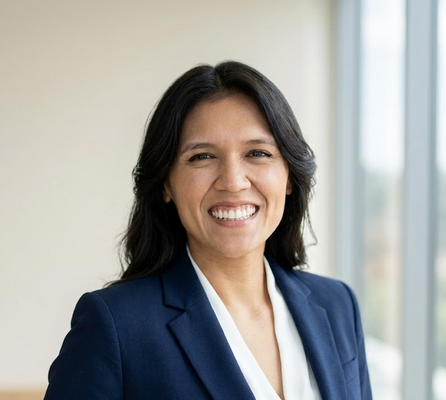 a medium shot of a woman with long, dark wavy hair smiling broadly at the camera. she is wearing a navy blue  blazer over a white V-neck blouse, set against a bright, softly blurred office background.