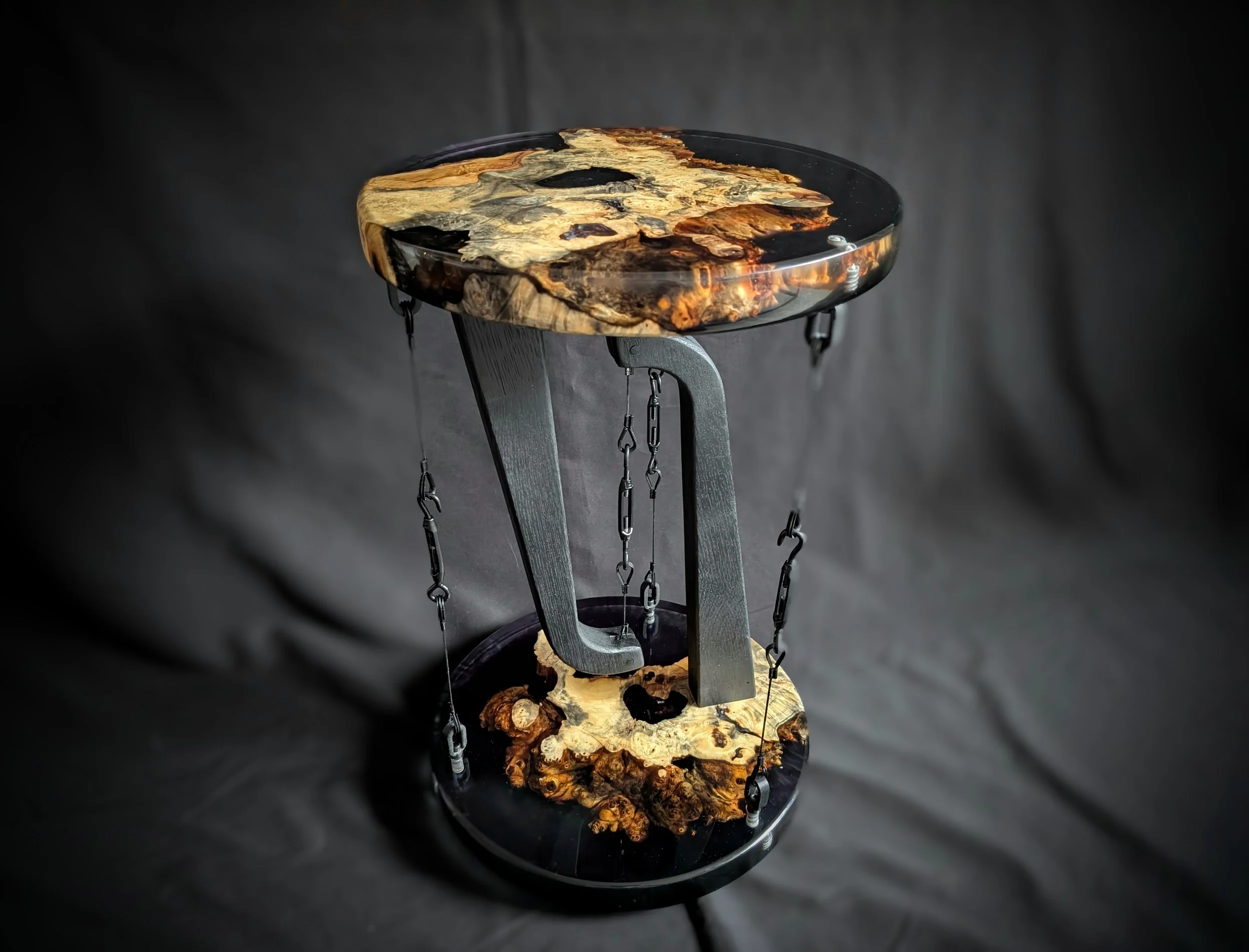 A decorative table with a circular top and bottom made of polished wood with natural knots and grain, supported by a black metal frame with chains for support, set against a dark background.