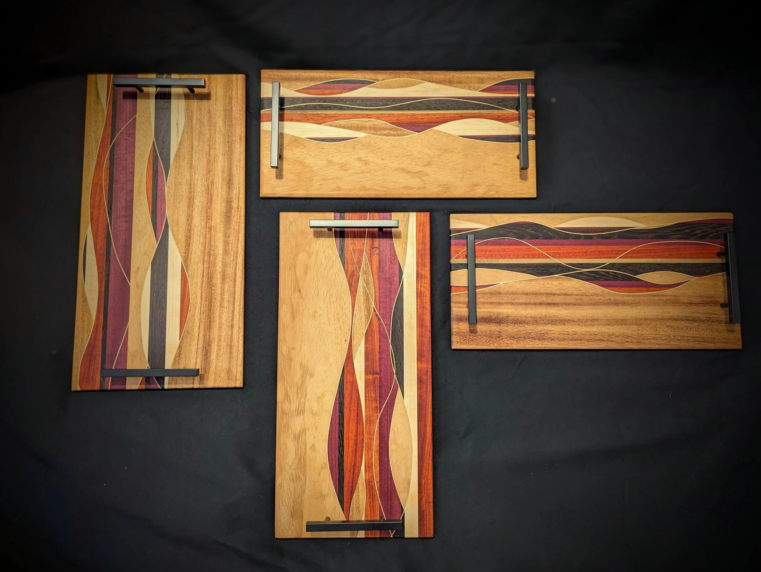 Four decorative wooden cutting boards with abstract paint designs and metal handles, arranged on a black surface.