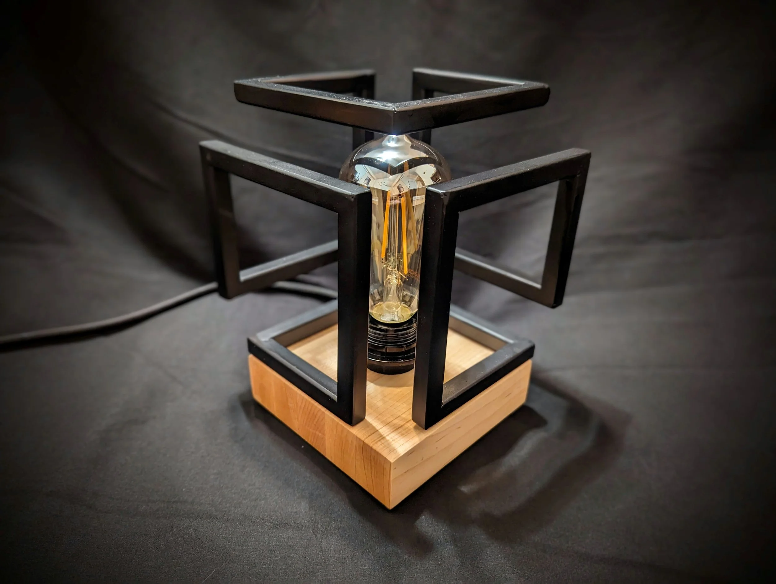 A decorative table lamp with a wooden base, a central glass bulb, and a geometric black metal frame surrounding the bulb.