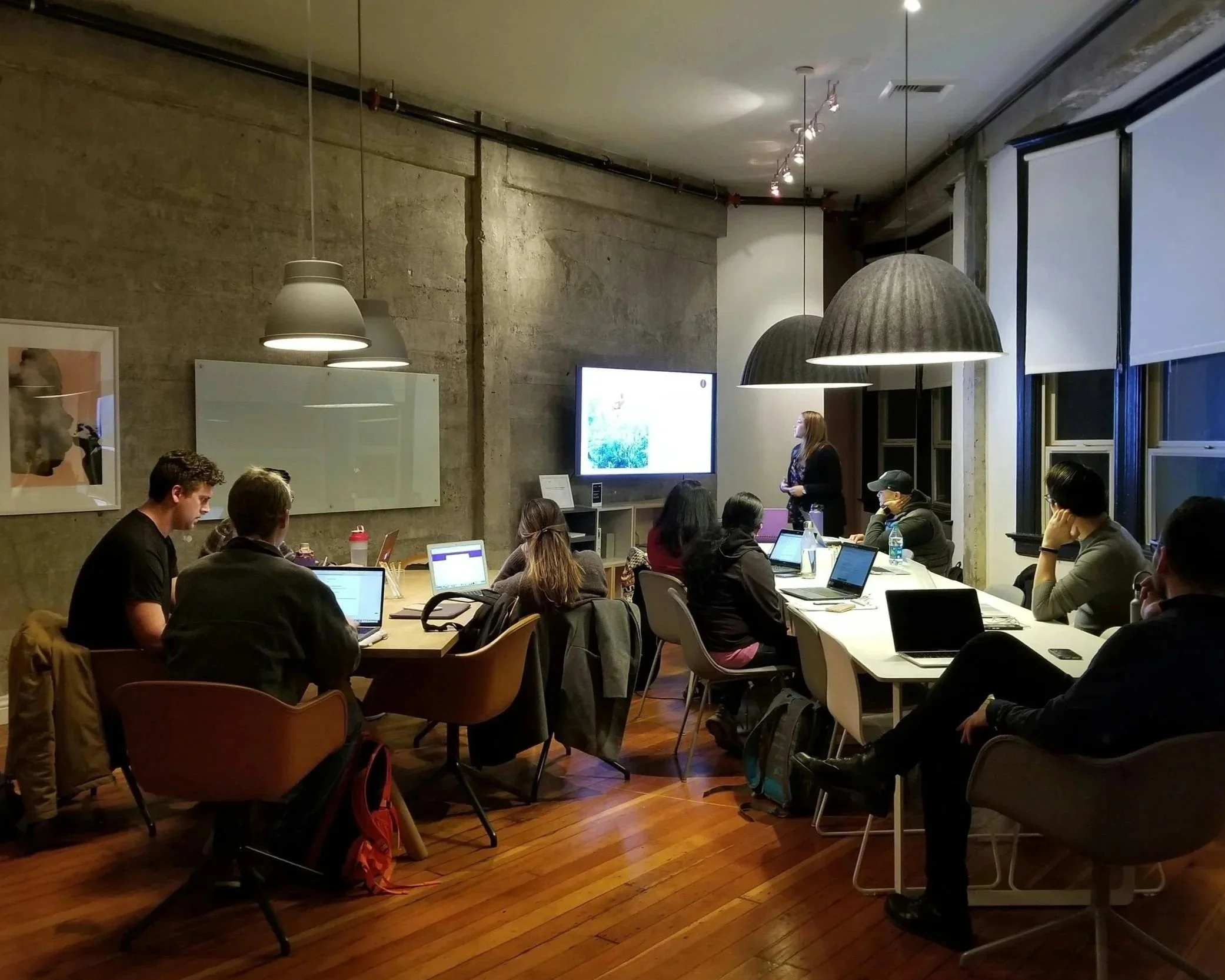 A group of people attending a meeting in a conference room, sitting at tables with laptops, facing a woman presenting in front of a large screen.