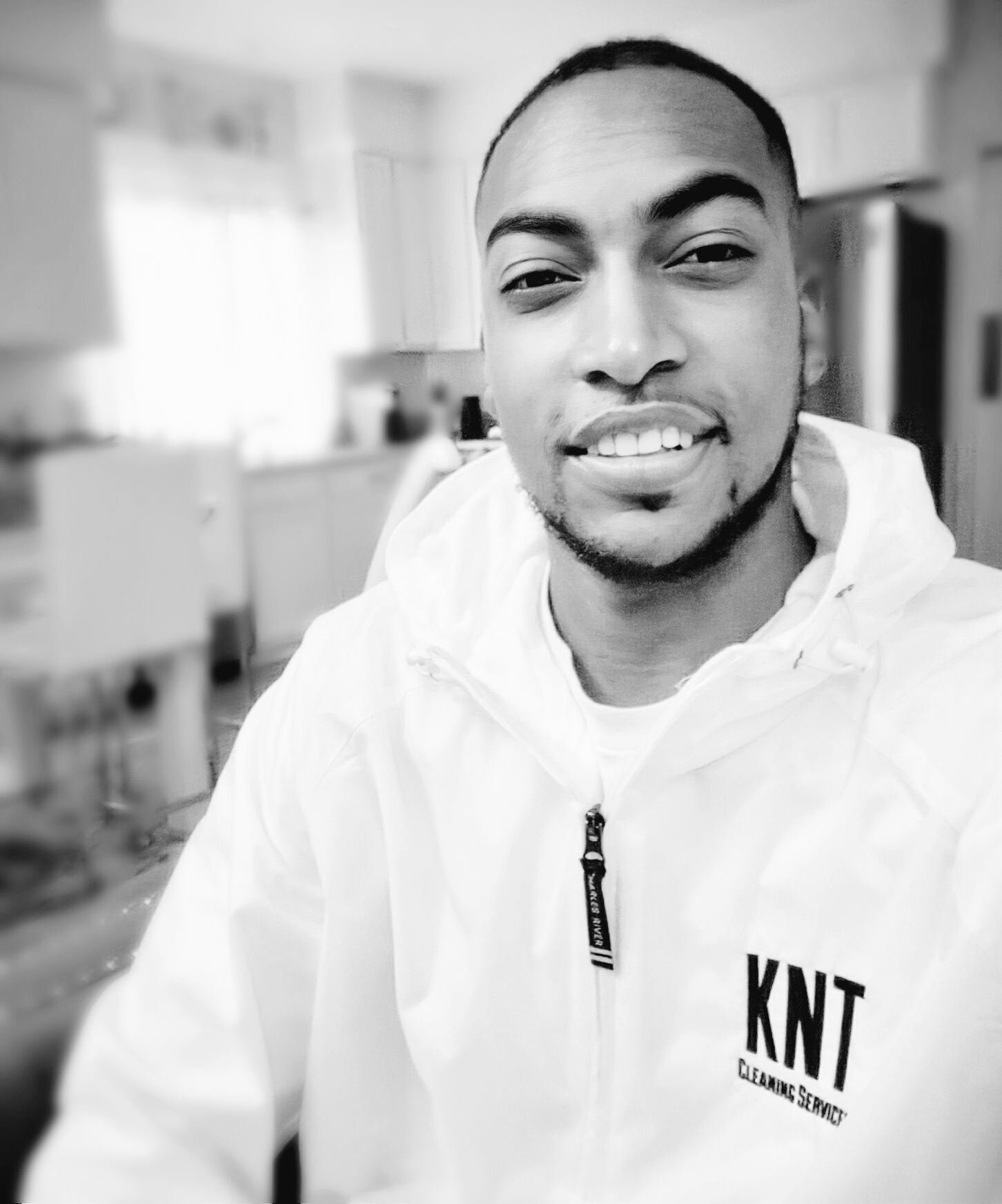 A smiling man wearing a white jacket with the logo KNT Cleaning Service, standing in a kitchen.