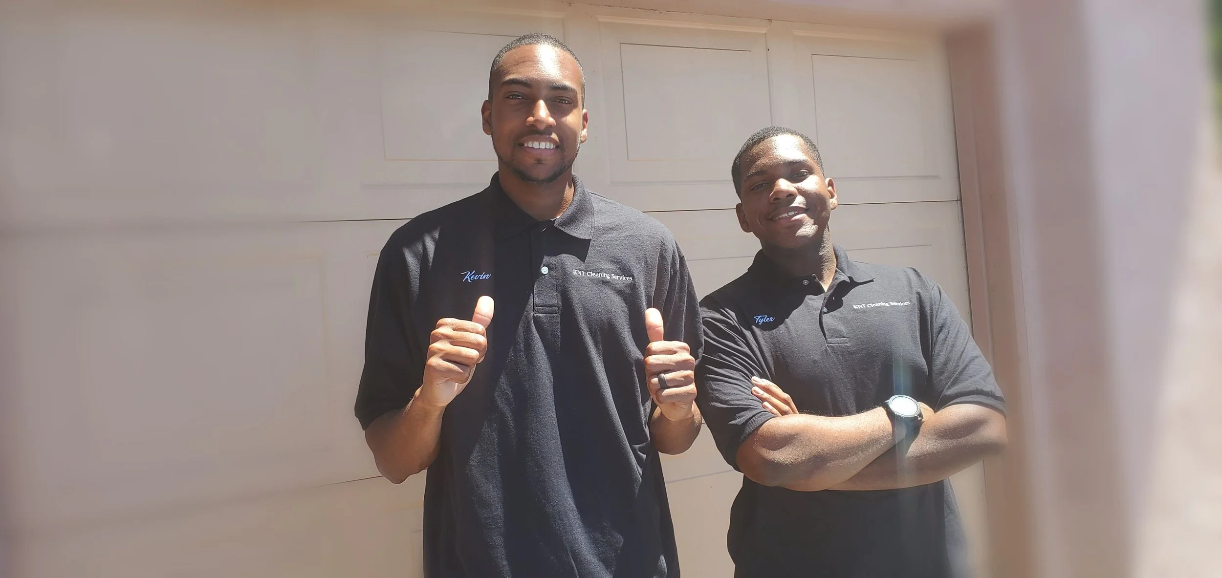 Two men wearing black polo shirts with customized names and company logo, standing in front of a beige garage door, smiling at the camera.