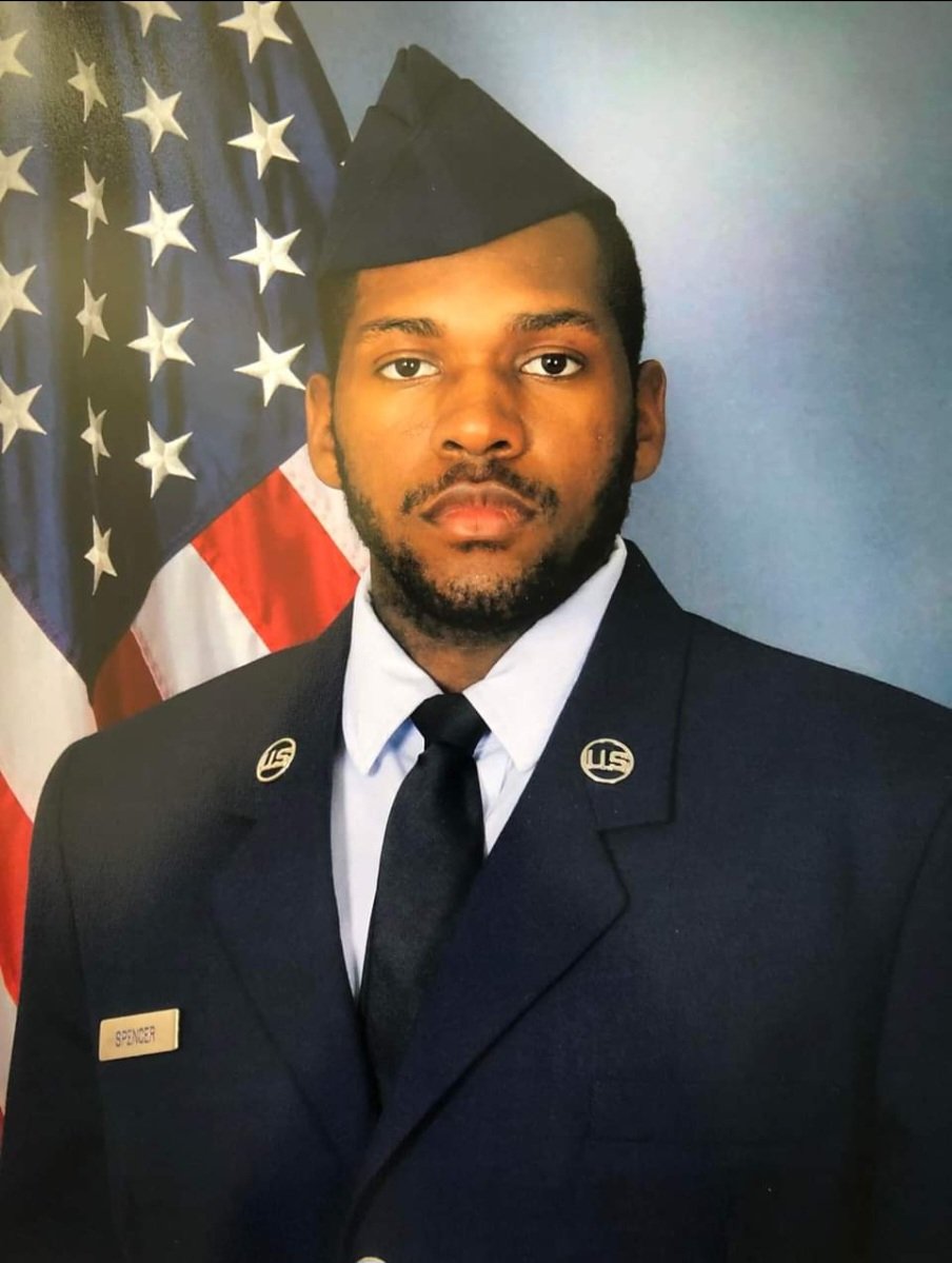 A man in a military dress uniform with a name tag 'Spencer', standing in front of United States flags.
