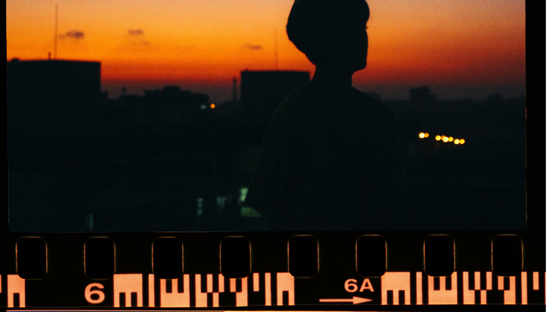 Silhouette of a person with short hair and glasses standing outdoors during sunset, with a cityscape in the background.