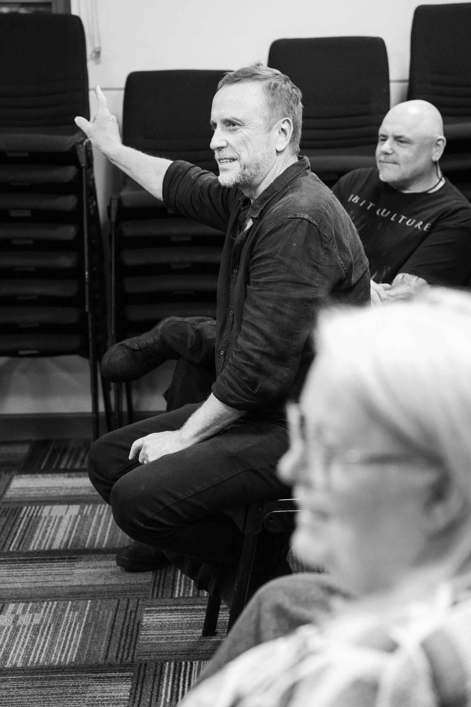A black and white photo of a man sitting on a chair, in profile, gesturing with his right hand, with other people in the background.