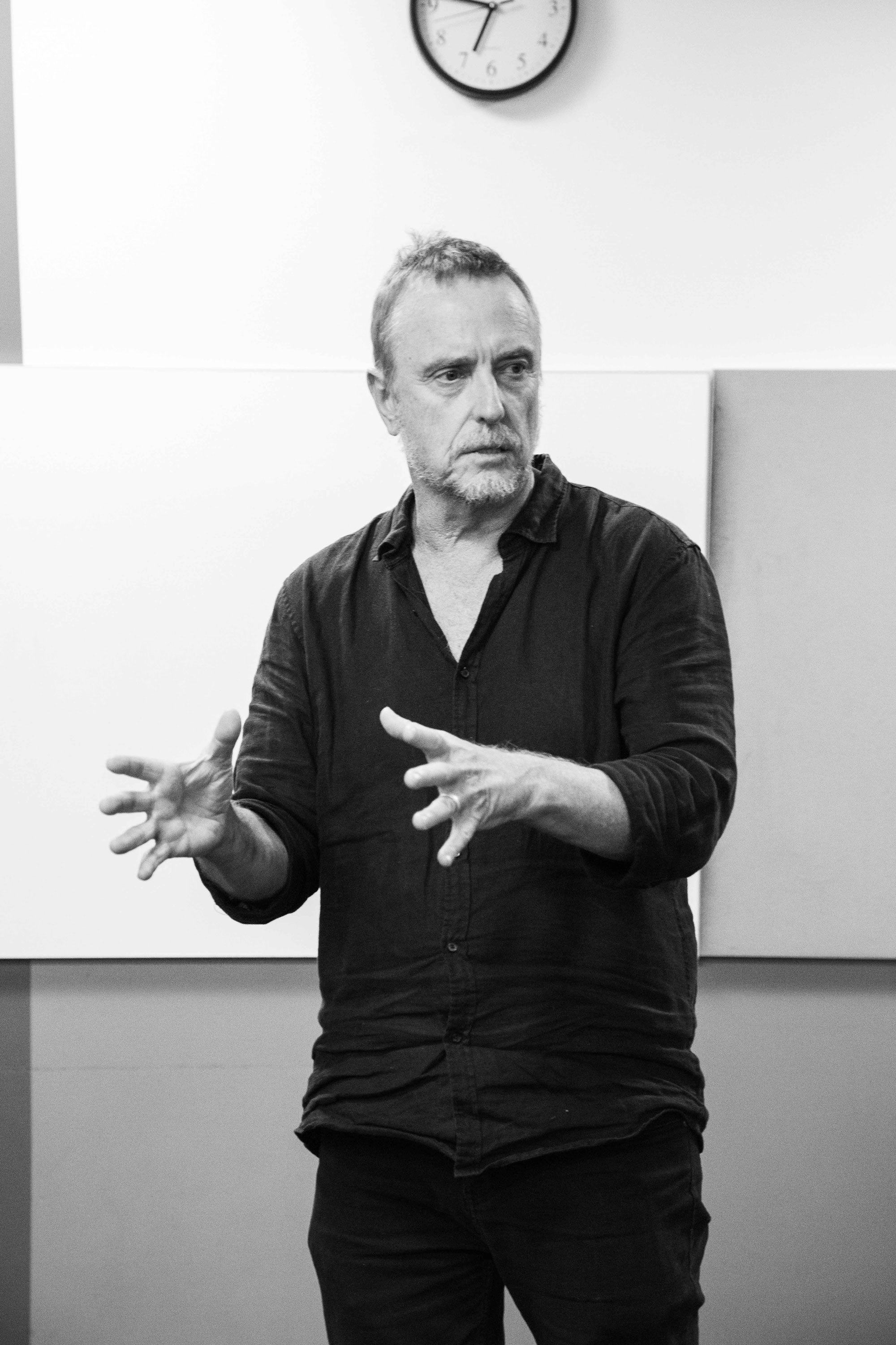 A man in a dark shirt gestures with his hands while speaking or explaining in a room with a clock on the wall.