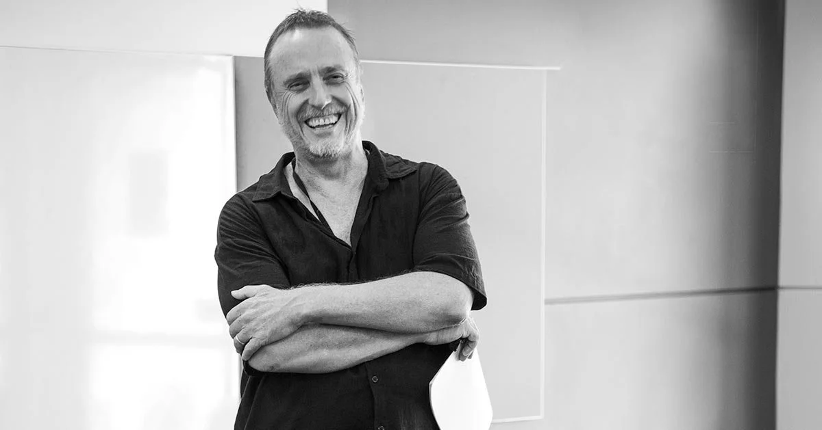 Black and white photo of a man with short hair, a beard, and a mustache, smiling with arms crossed, standing indoors against a plain background.