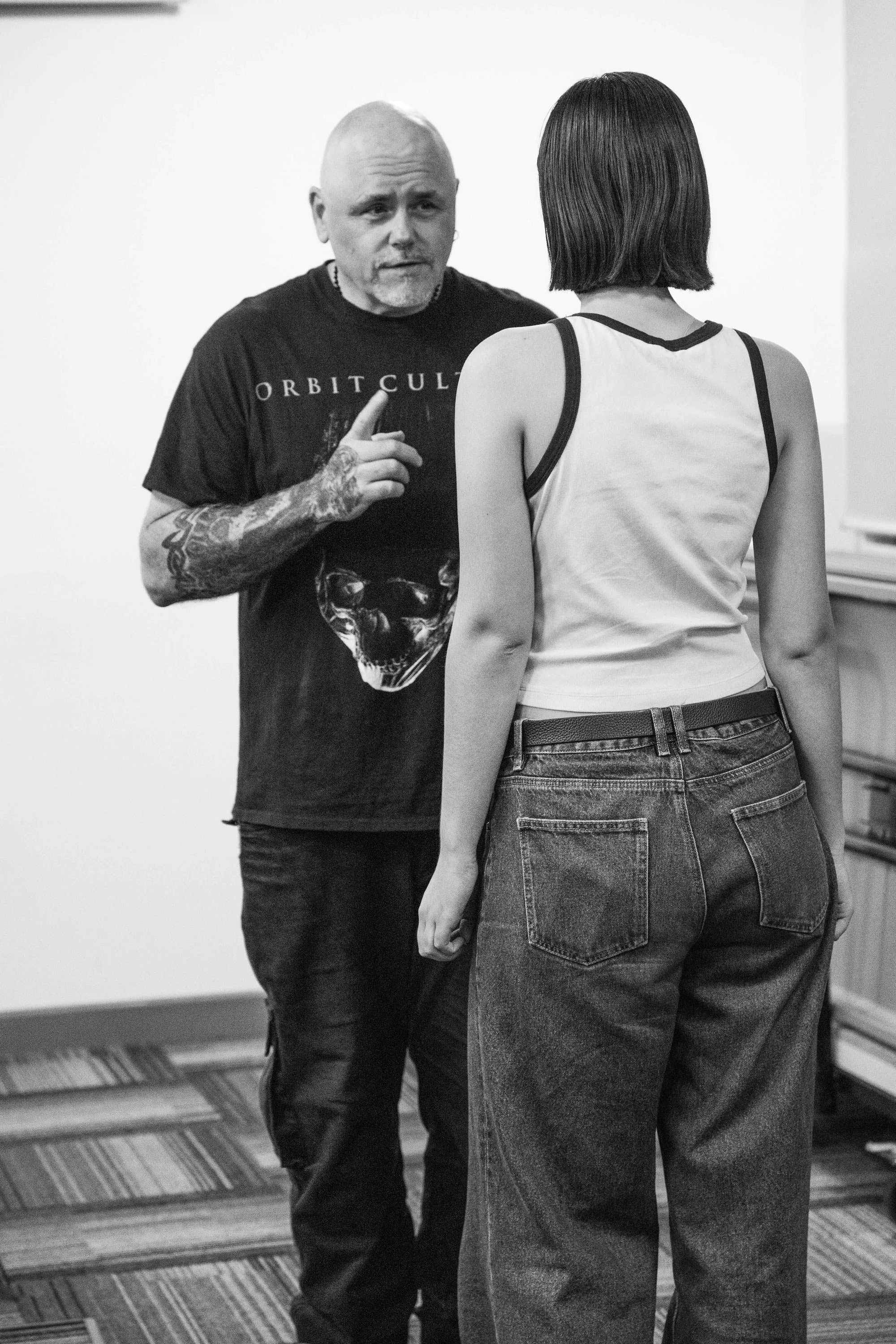 A man with tattoos on his arm and a bald head is talking to a woman with short hair. The man is wearing a black T-shirt with a skull graphic and the words 'Orbit Cult'. The woman is wearing a white tank top and high-waisted jeans, standing with her b