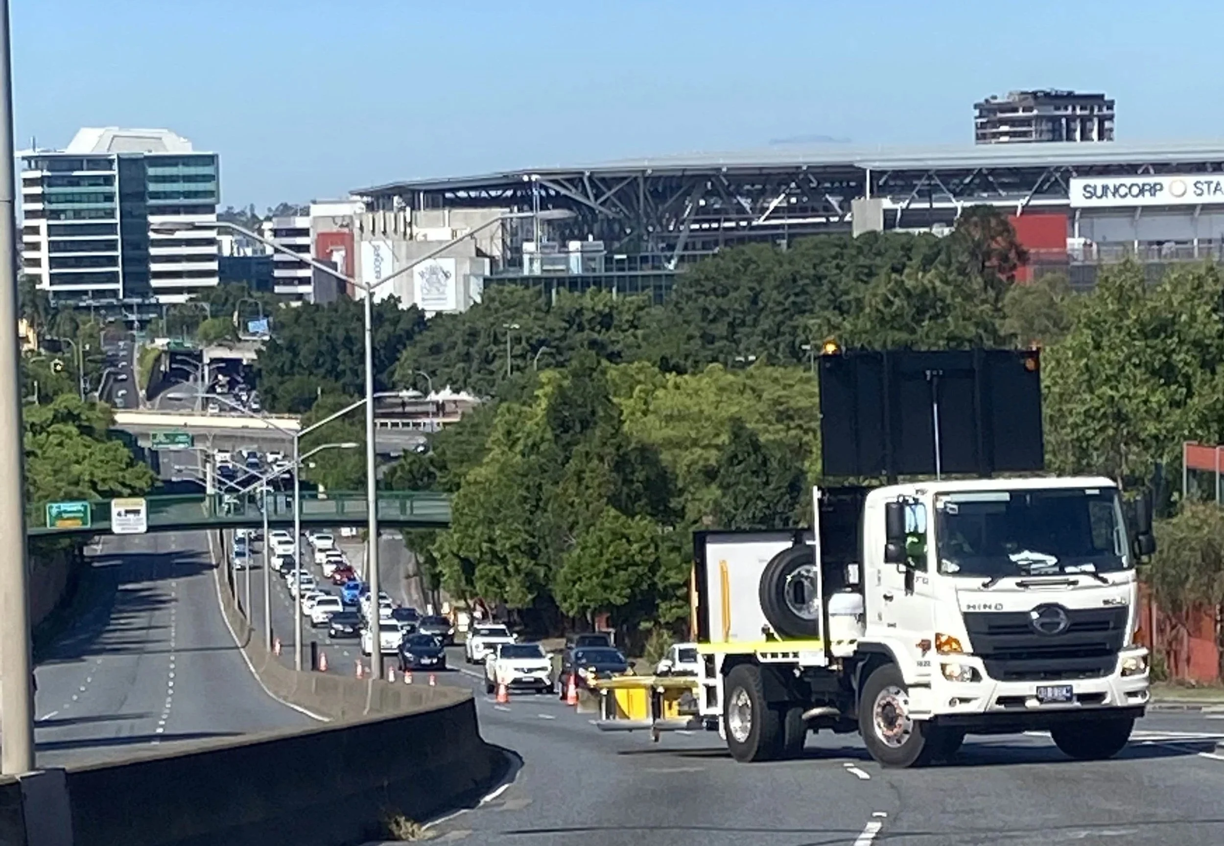 A TMA blocking off a highway. Traffic cones are placed along the road. In the background, there are trees and modern buildings, including a stadium with a sign that says 'Suncorp Stadium'.