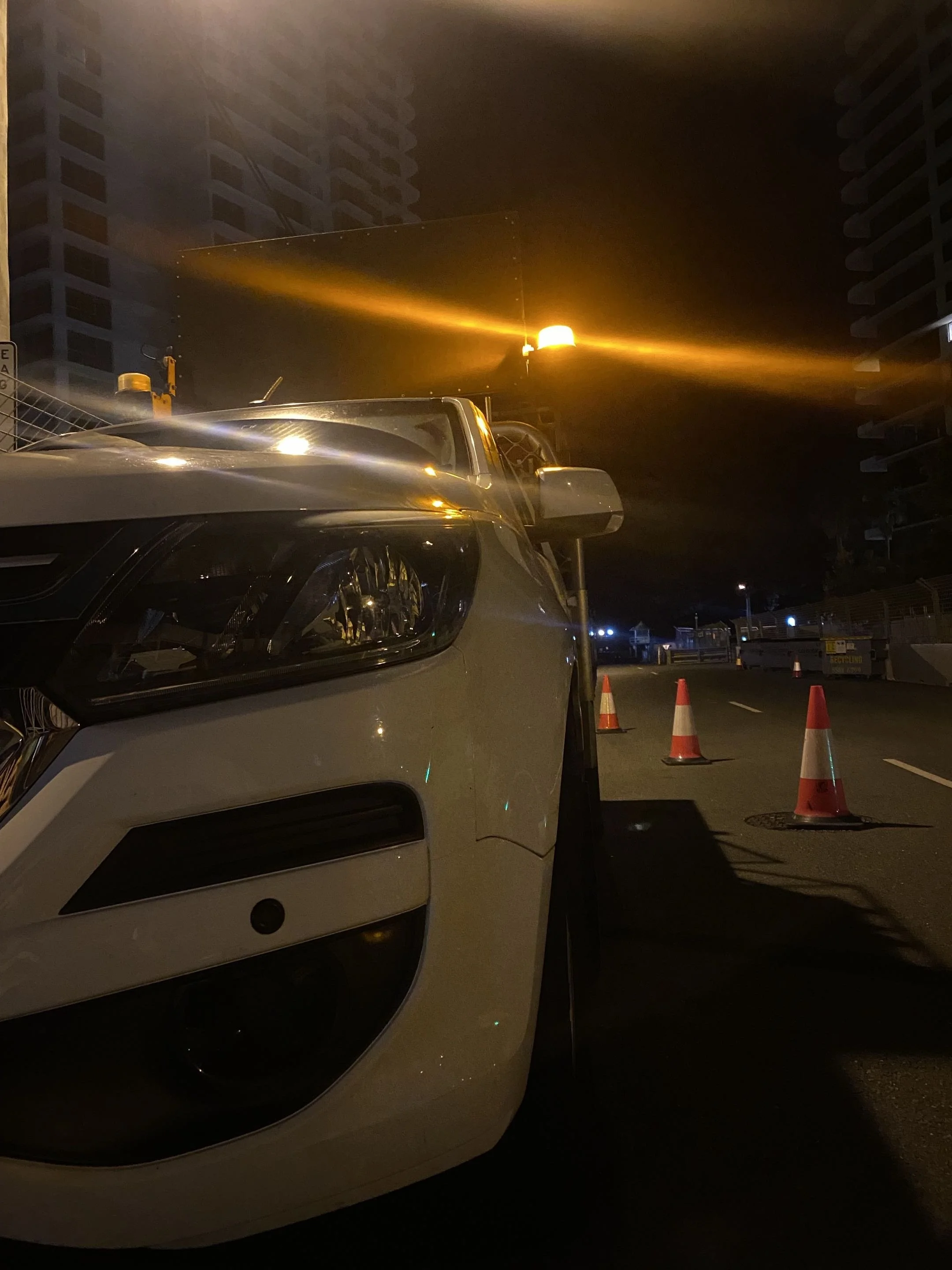 A parking lot at night with orange traffic cones, a white vehicle in the foreground, and tall buildings in the background; a streetlight illuminates part of the scene.