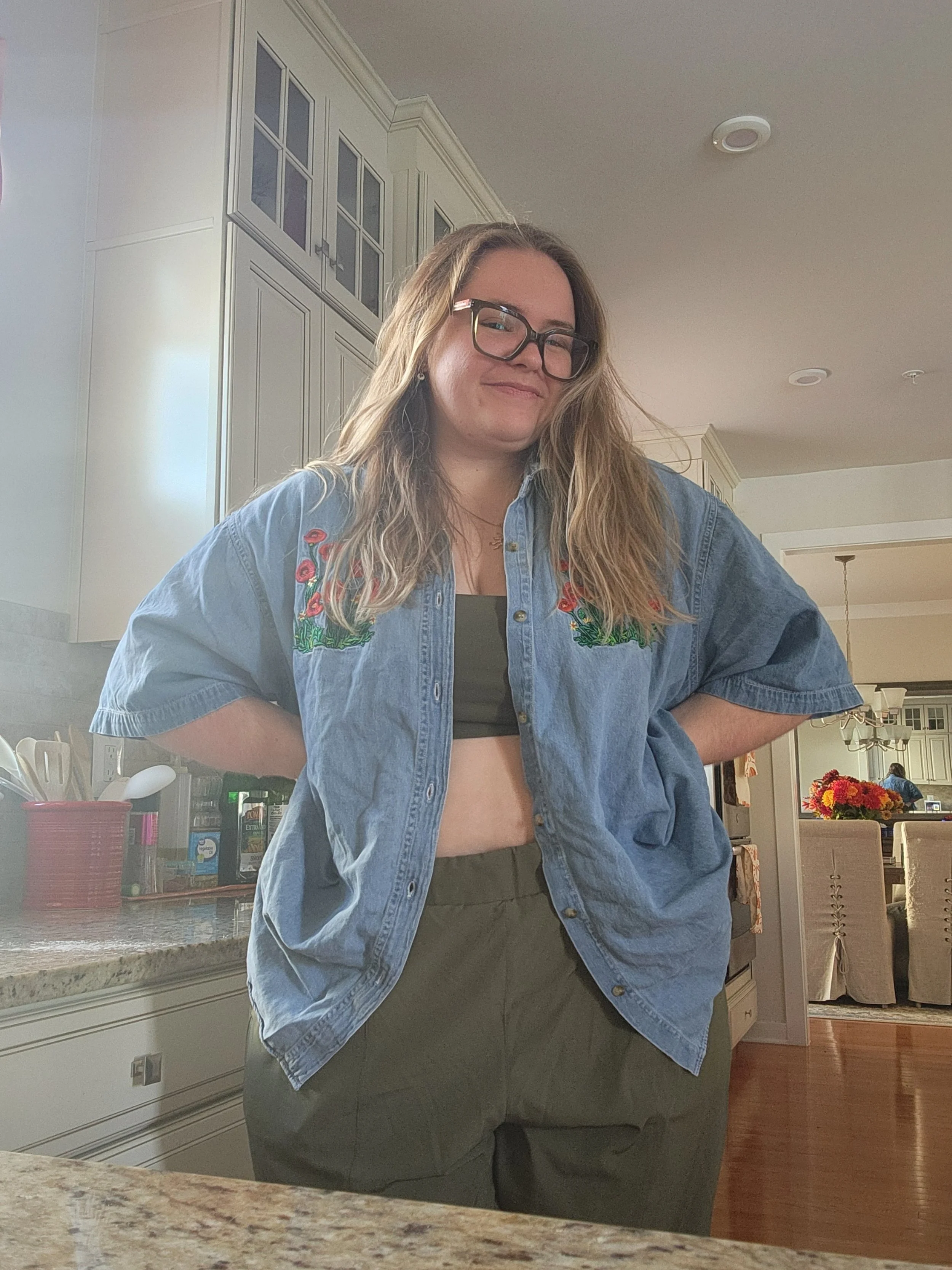 A woman with glasses and wavy blonde hair standing in a kitchen, smiling with hands on hips, wearing a denim shirt over a black top and green pants.