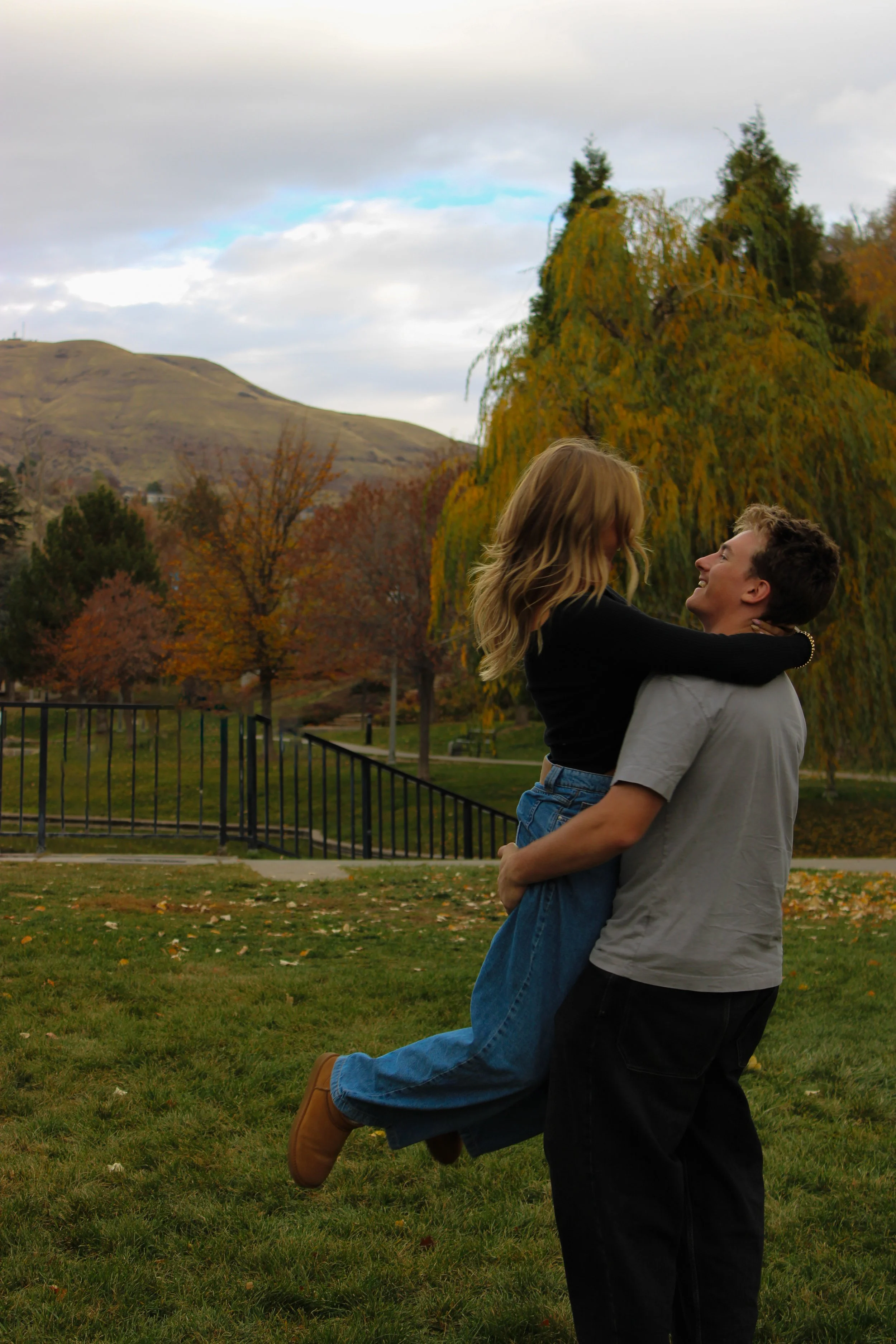A young couple enjoying a playful moment outdoors in a park during fall, with colorful trees and a hill in the background.