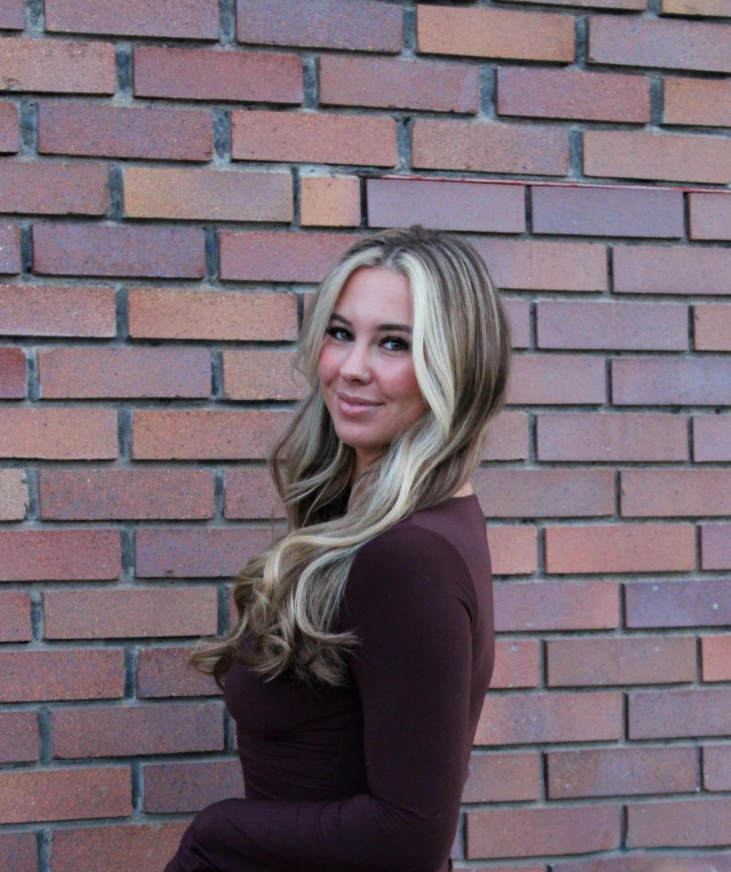 A woman with long, wavy blonde hair and a nose piercing standing in front of a brick wall, smiling slightly at the camera.
