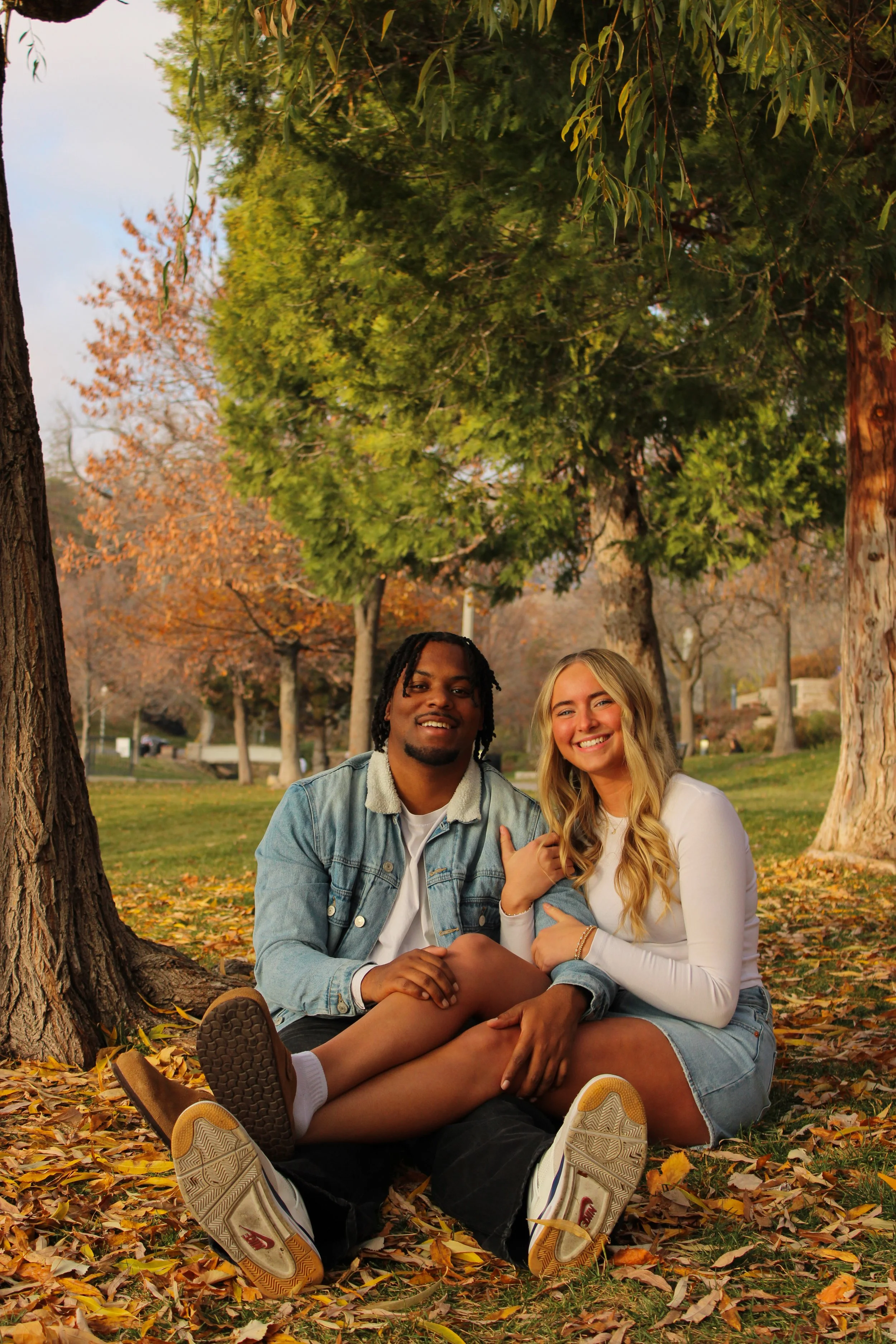 A young couple sitting under trees with fall foliage, smiling at the camera.