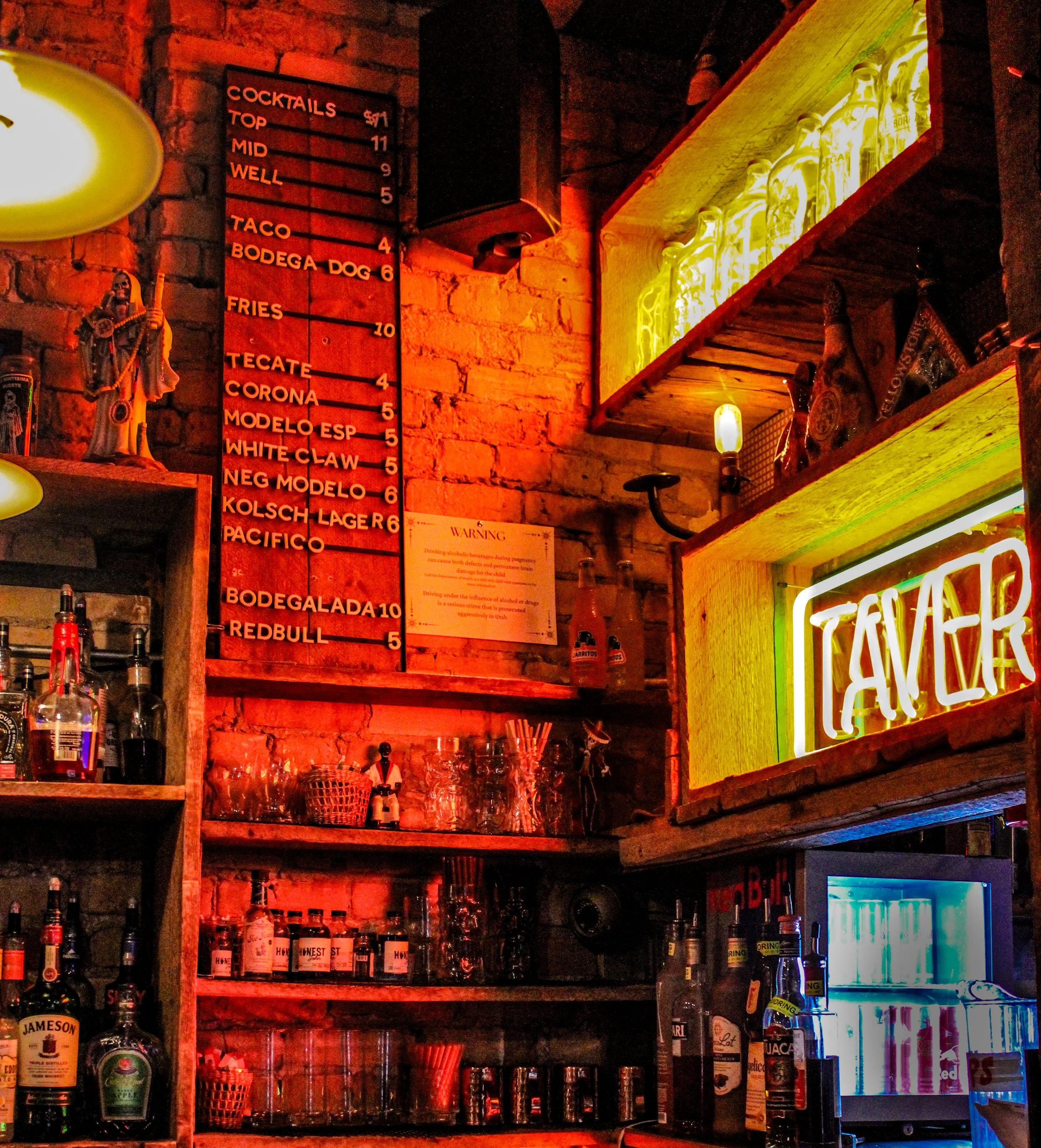 A bar counter area with a brick wall, displaying a black and red menu board listing drinks and their prices, illuminated by orange and yellow lighting. Shelves hold liquor bottles, glassware, and decorative items, including a statue and neon sign rea