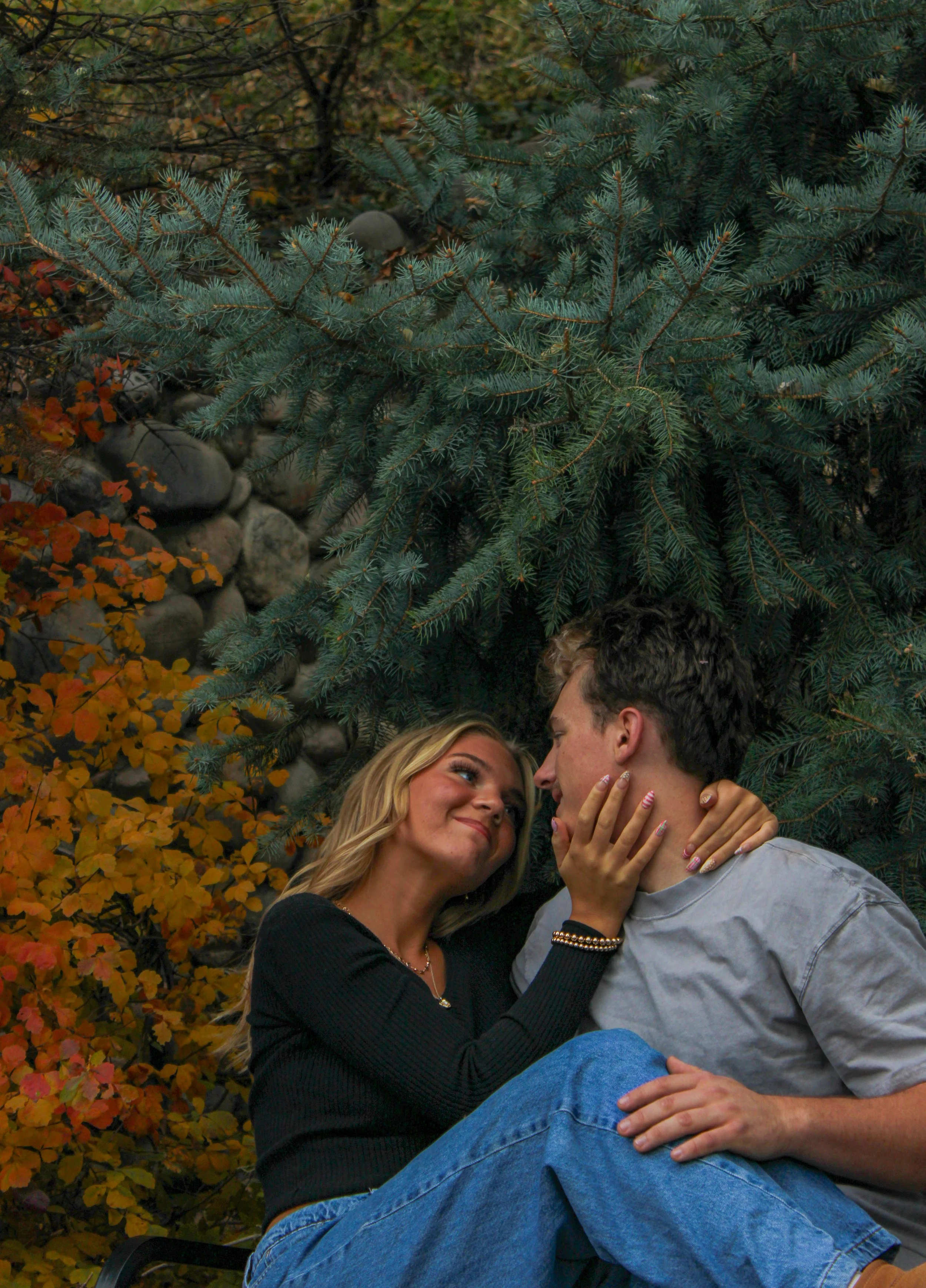 A young couple sitting close together outdoors in front of a pine tree and fall foliage, gazing into each other's eyes with smiles.