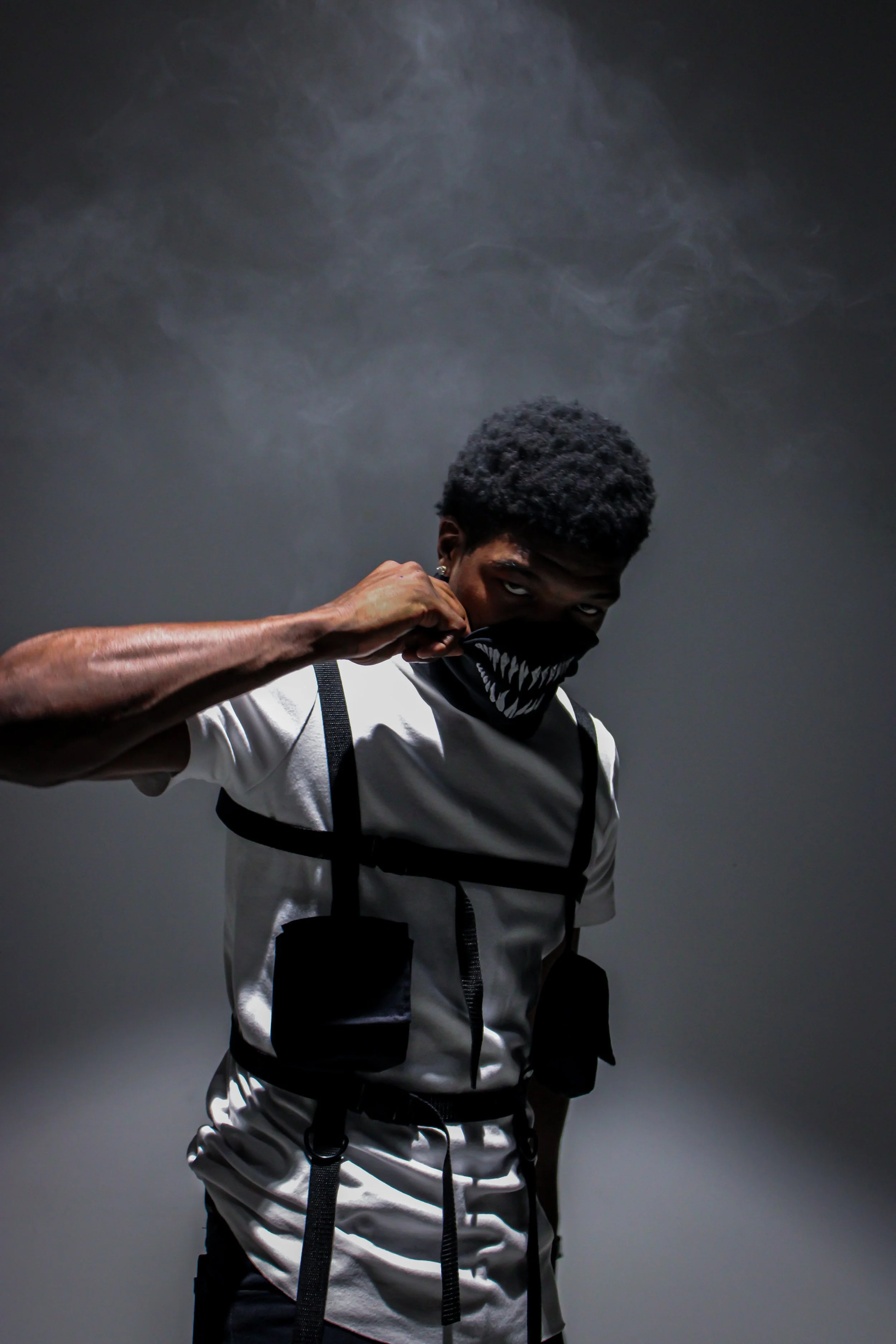 A young man with a dark hoodie with a sinister face mouth design, standing in front of a smoky gray background, pulling down his mask to reveal his eyes.