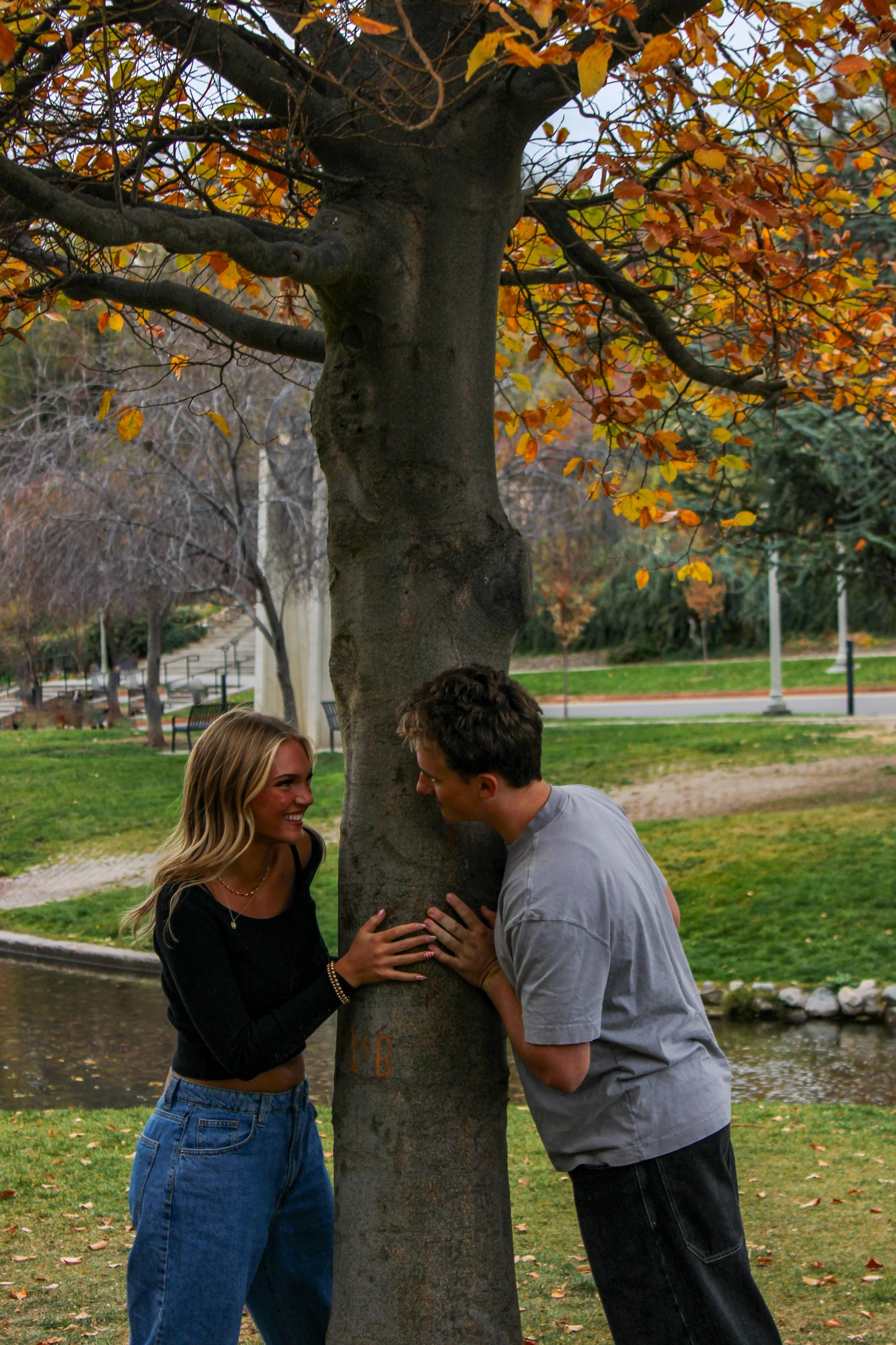 A young woman and young man smiling and touching a large tree trunk in a park during autumn.
