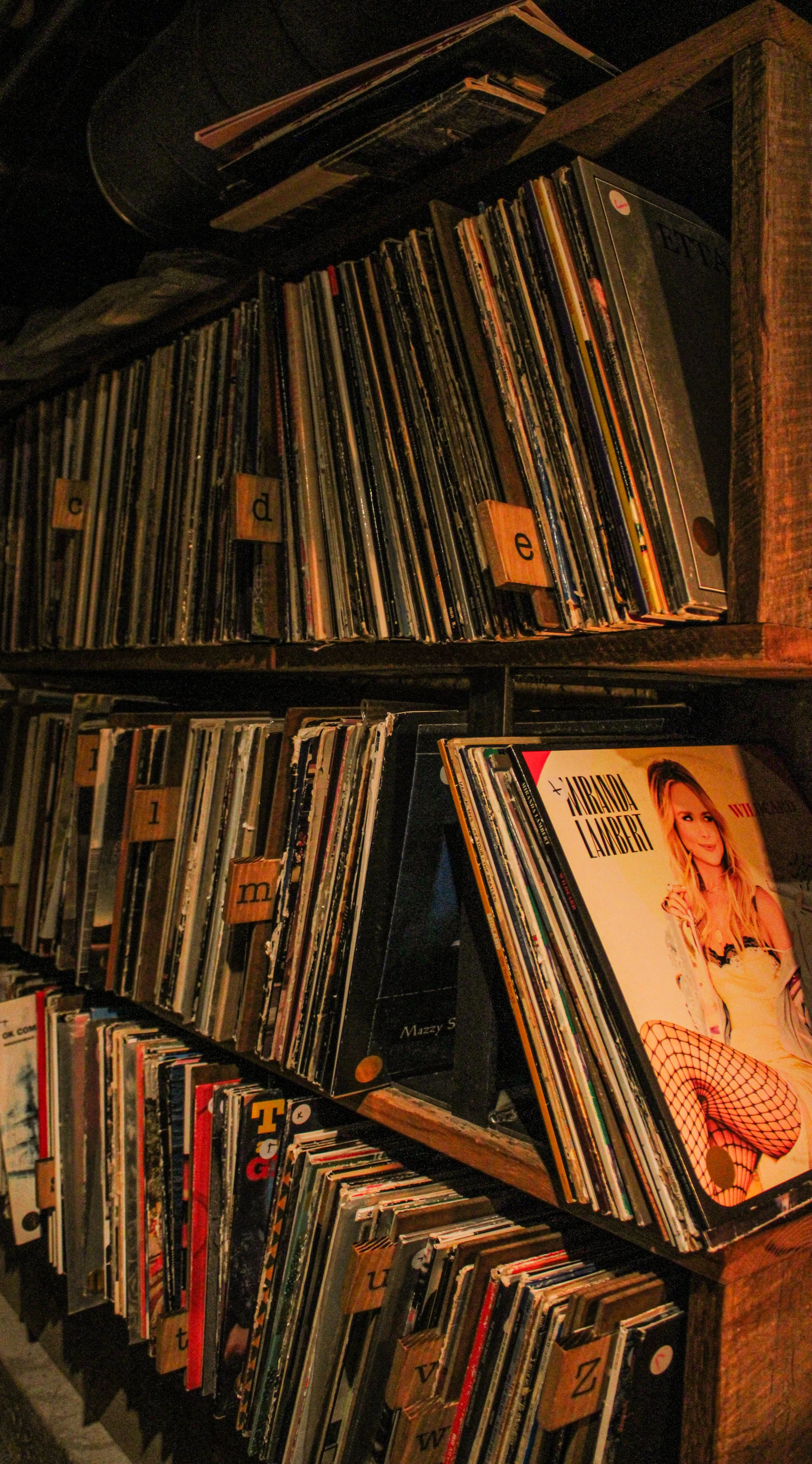 A wooden shelf filled with vinyl records organized alphabetically, with some categories labeled with letter tags, and a visible record album cover featuring Miranda Lambert.