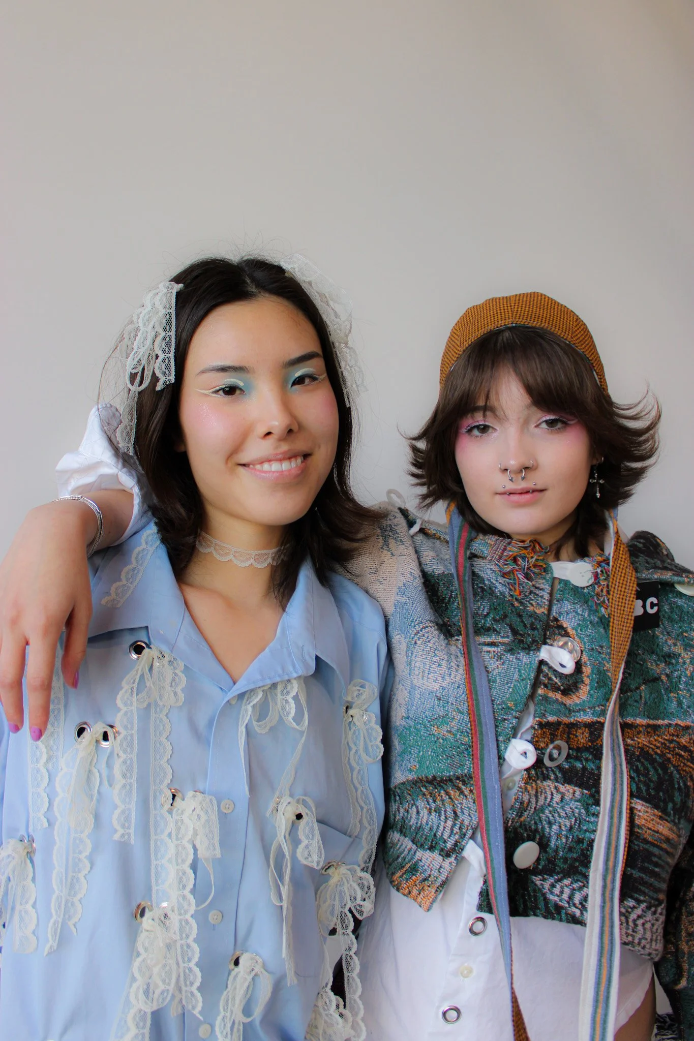 Two young women standing close, smiling at the camera, one with a lace headband and the other wearing a yellow cap, dressed in eclectic, colorful clothing with unique makeup.