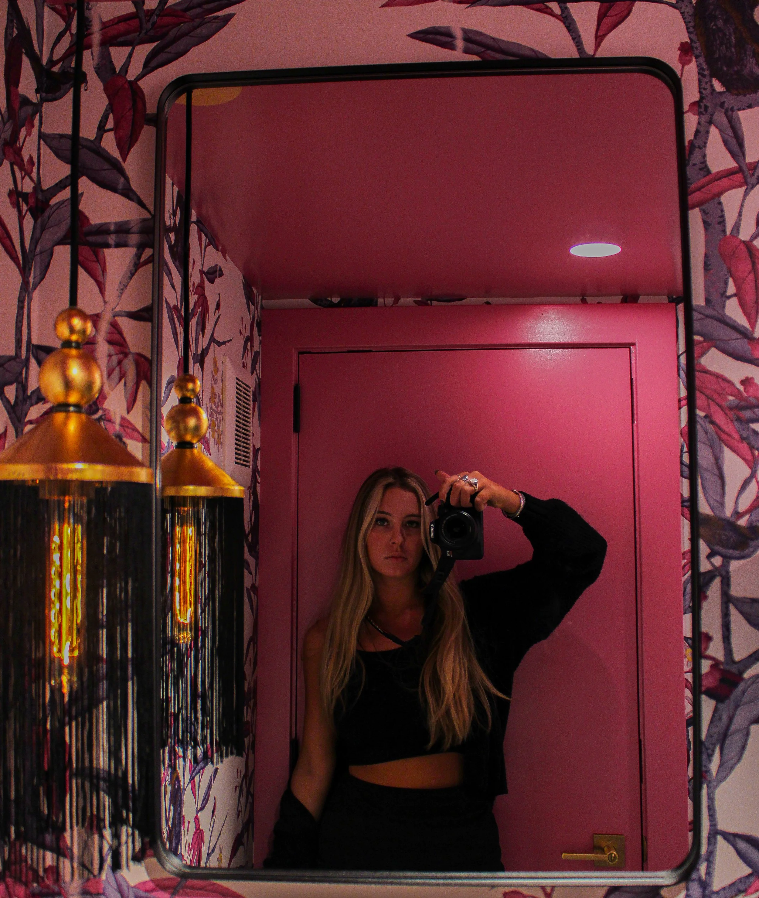 A young woman with long blonde hair taking a mirror selfie in a pink room with floral wallpaper. She is wearing a black crop top and black pants, holding a camera, with a serious expression.