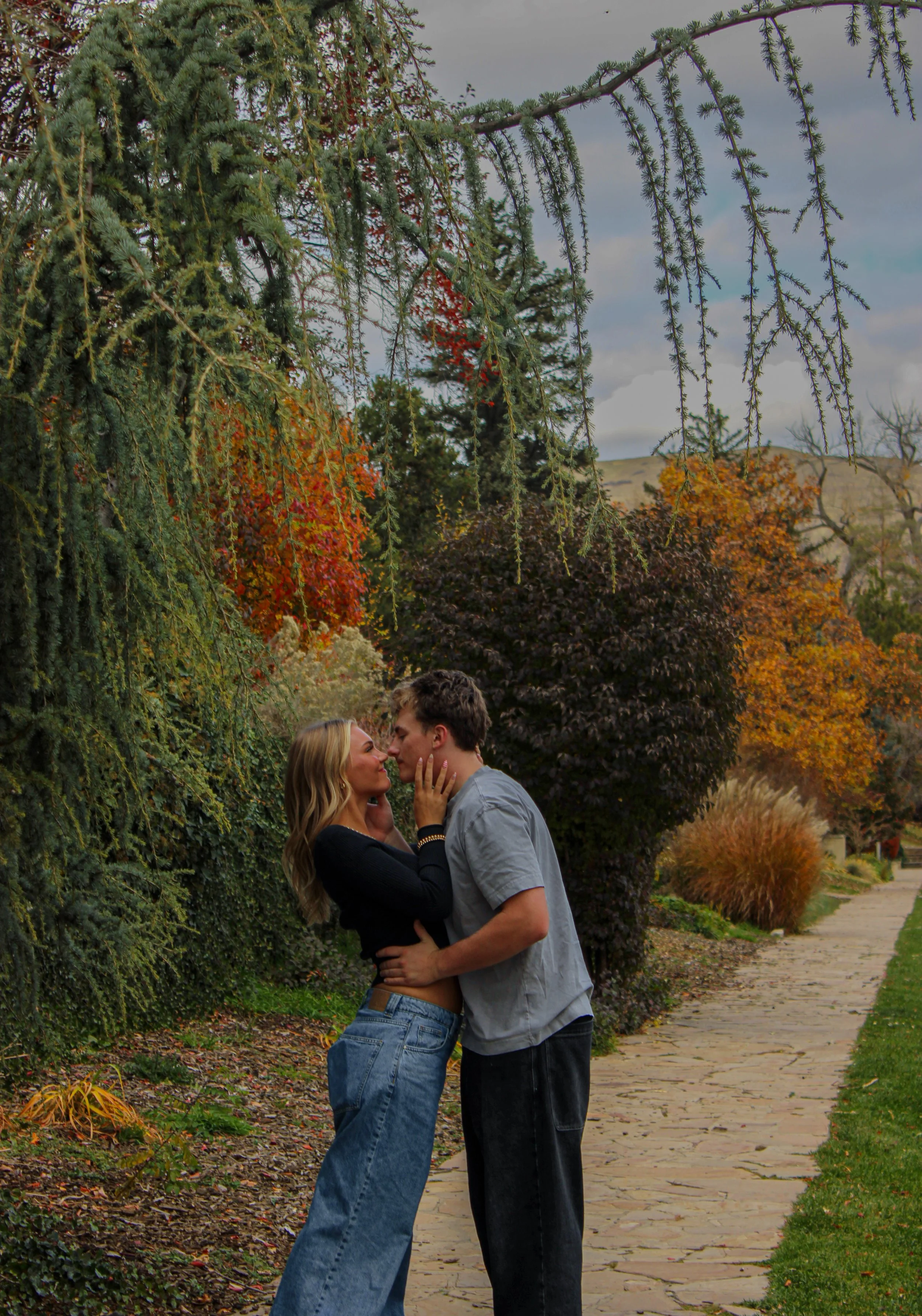 A couple embraces and nearly kisses on a stone pathway lined with fall foliage trees.