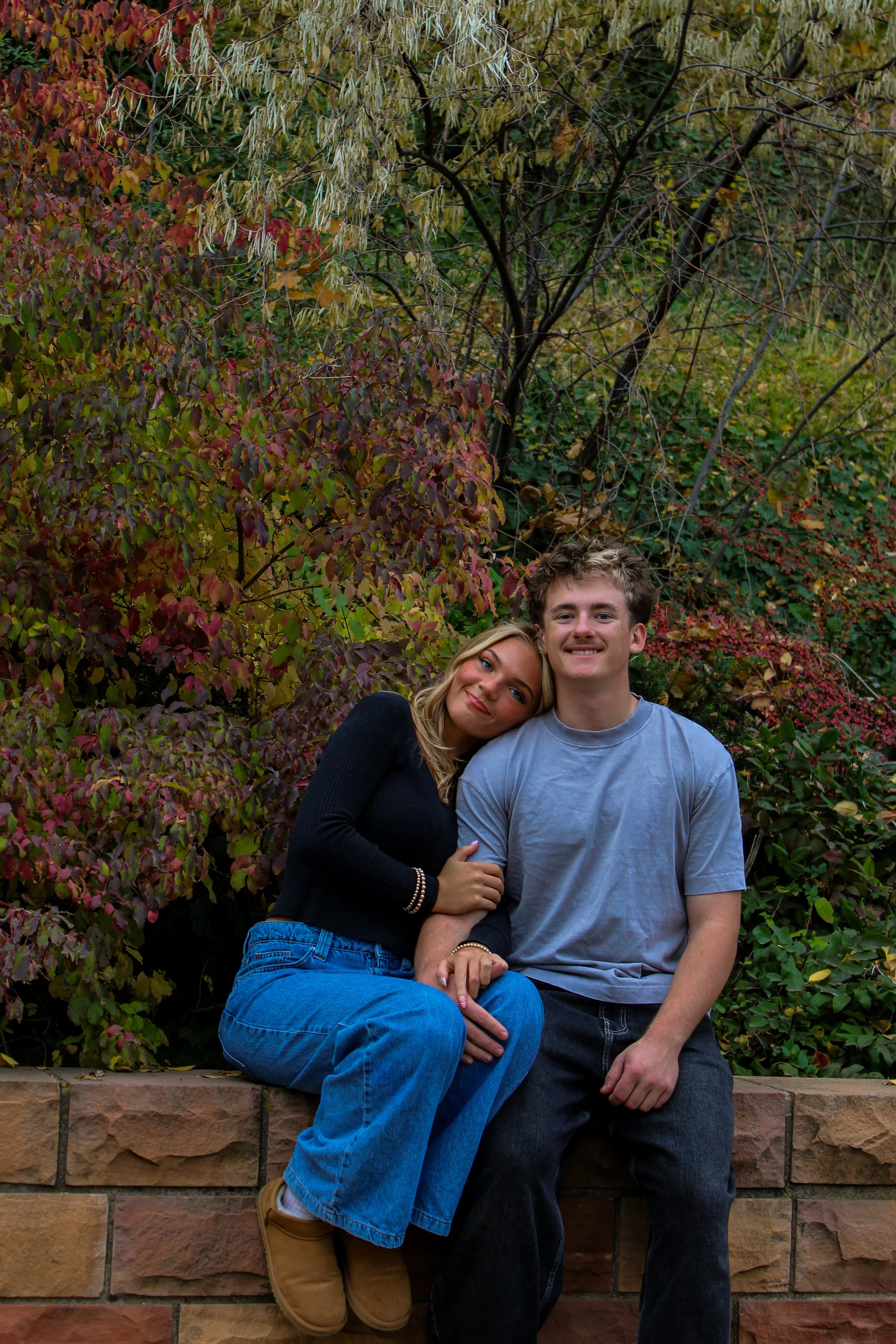 A young woman and a young man sitting together on a brick wall in a garden with fall foliage, the woman resting her head on the man's shoulder.