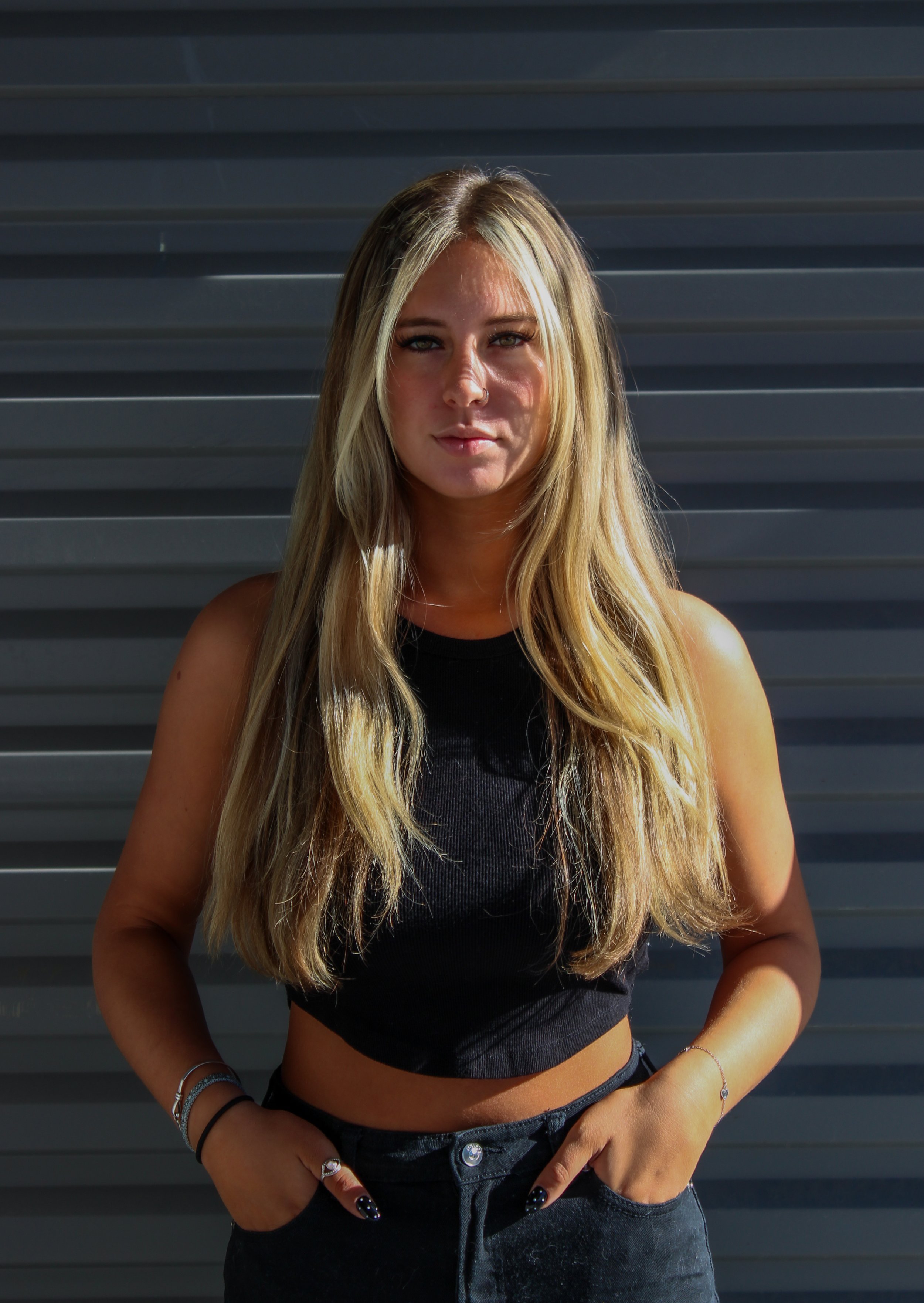 A young woman with long blonde hair, wearing a black sleeveless top and black jeans, stands with her hands in her pockets against a dark gray, horizontal metal siding background.