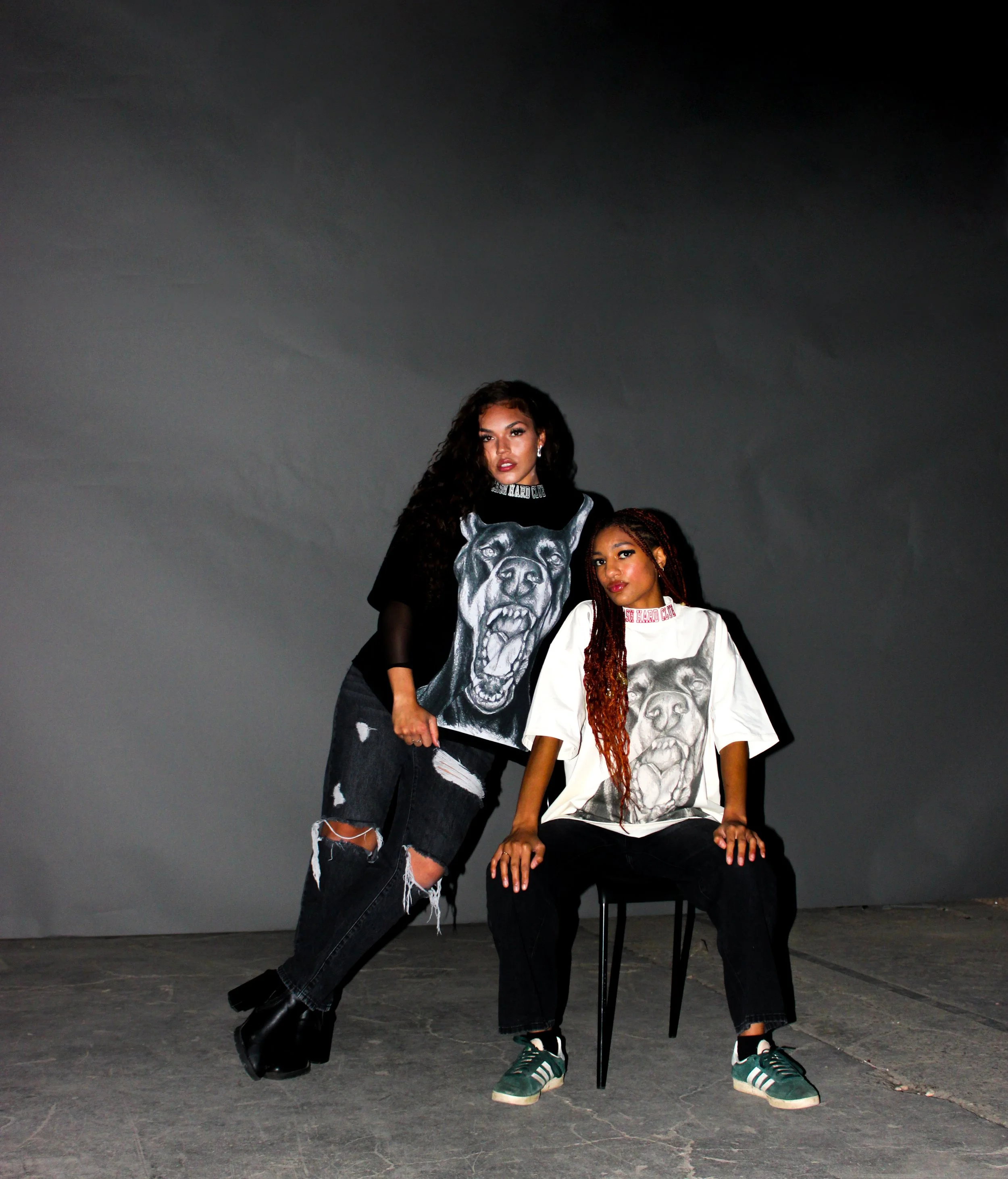 Two women posing against a dark gray background, wearing oversized T-shirts with a large dog illustration on the front.
