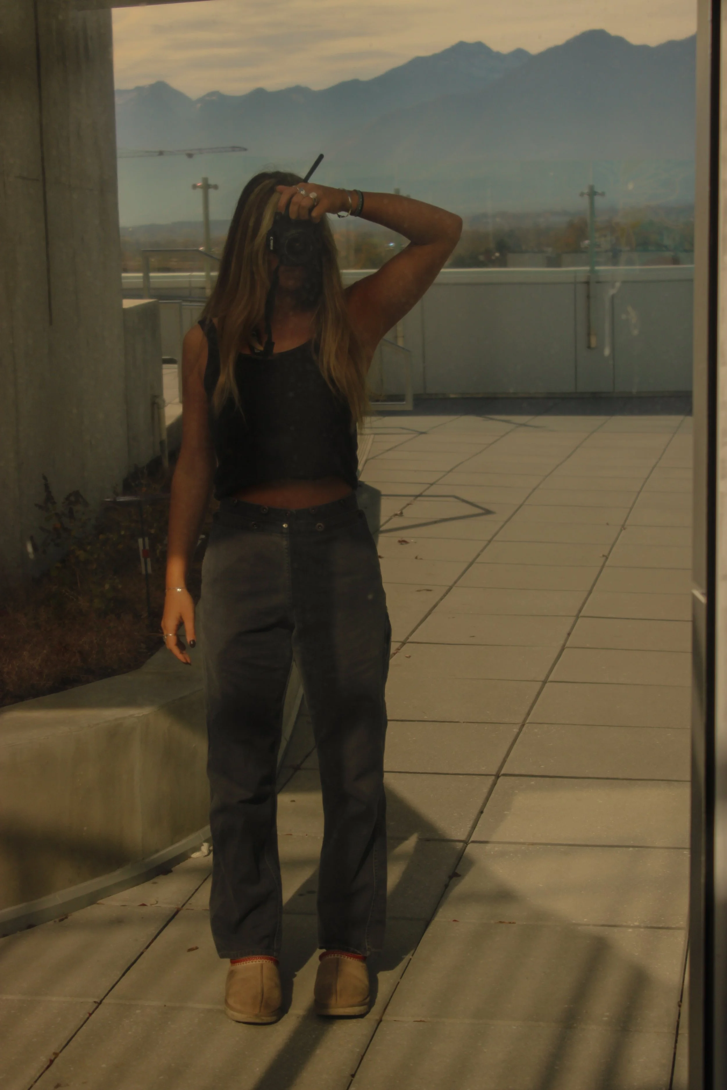 A woman taking a mirror selfie outdoors shows her reflection with mountains and a cloudy sky in the background. She is wearing a black sleeveless top, dark jeans, and tan shoes, and has long hair.