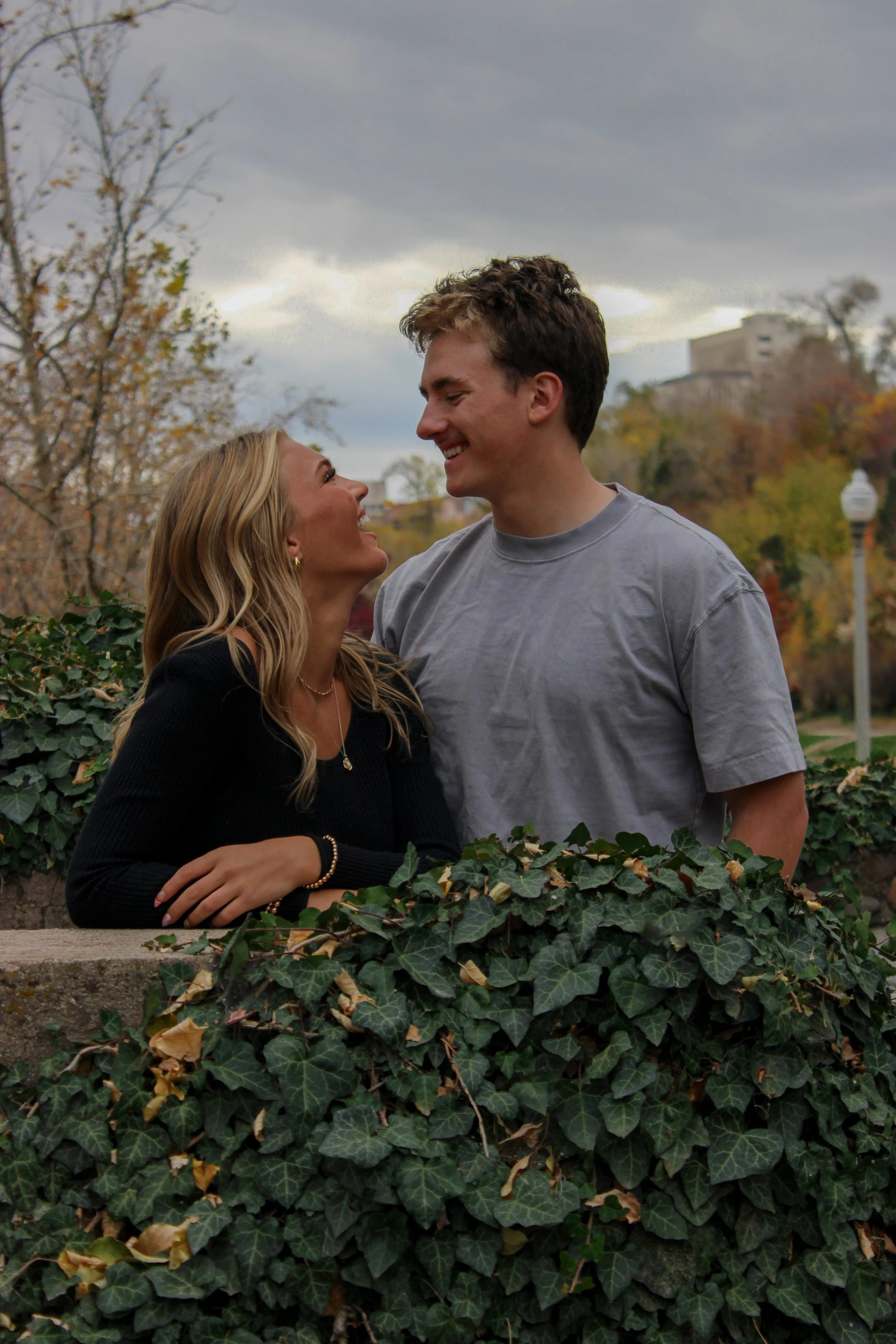 A young couple smiling and looking at each other, standing behind a hedge of green ivy in an outdoor park during autumn with trees showing fall colors and a cloudy sky.
