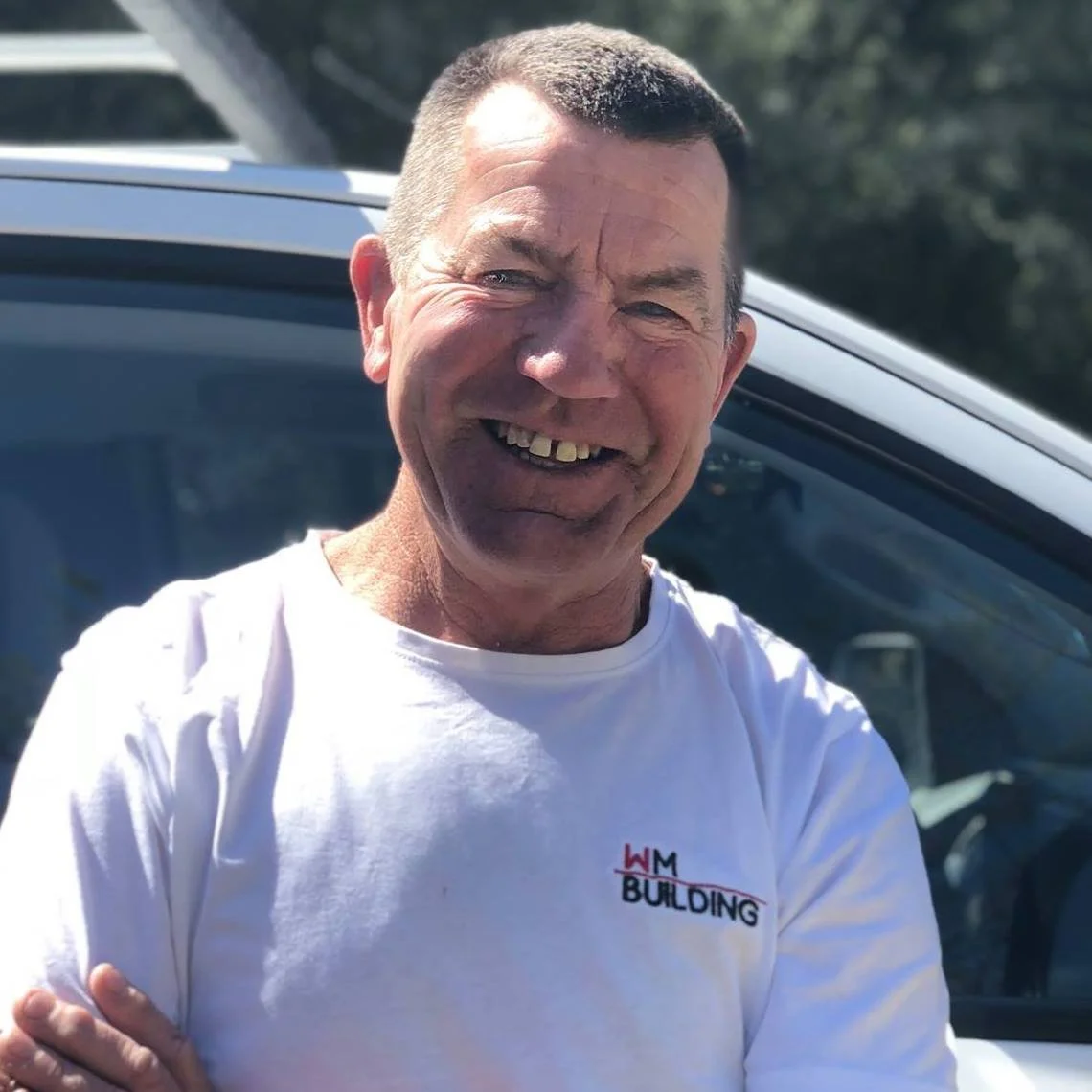 A smiling man with short dark hair and missing teeth, wearing a white T-shirt with 'H M BUILDING' logo, standing outdoors in front of a car.